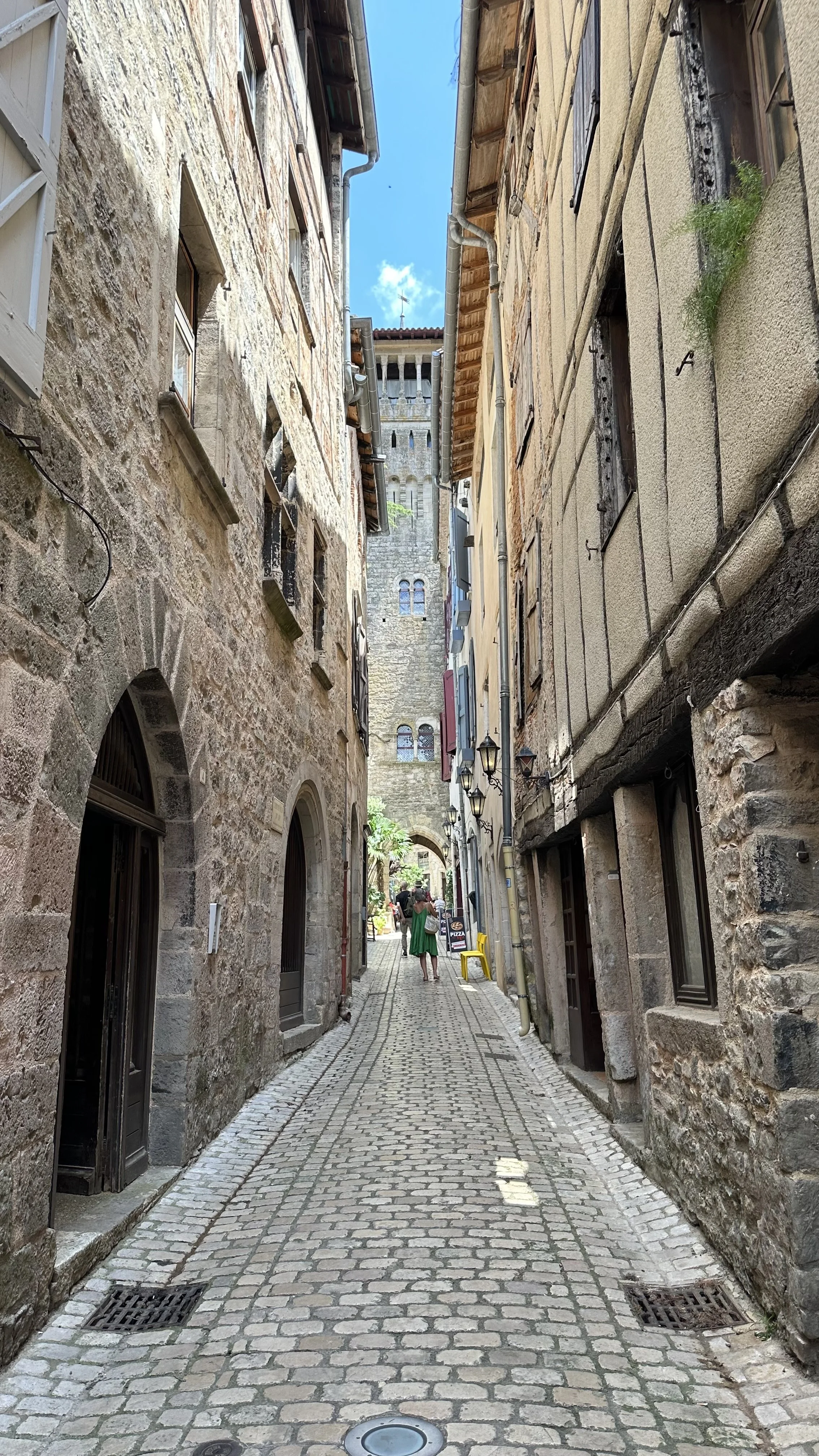 A narrow cobblestone street between old stone buildings with small windows, some with shutters, and a few hanging lanterns. There are a couple of people walking down the street and some outdoor seating at the end of the street. A church tower is visible in the background under a partly cloudy sky.