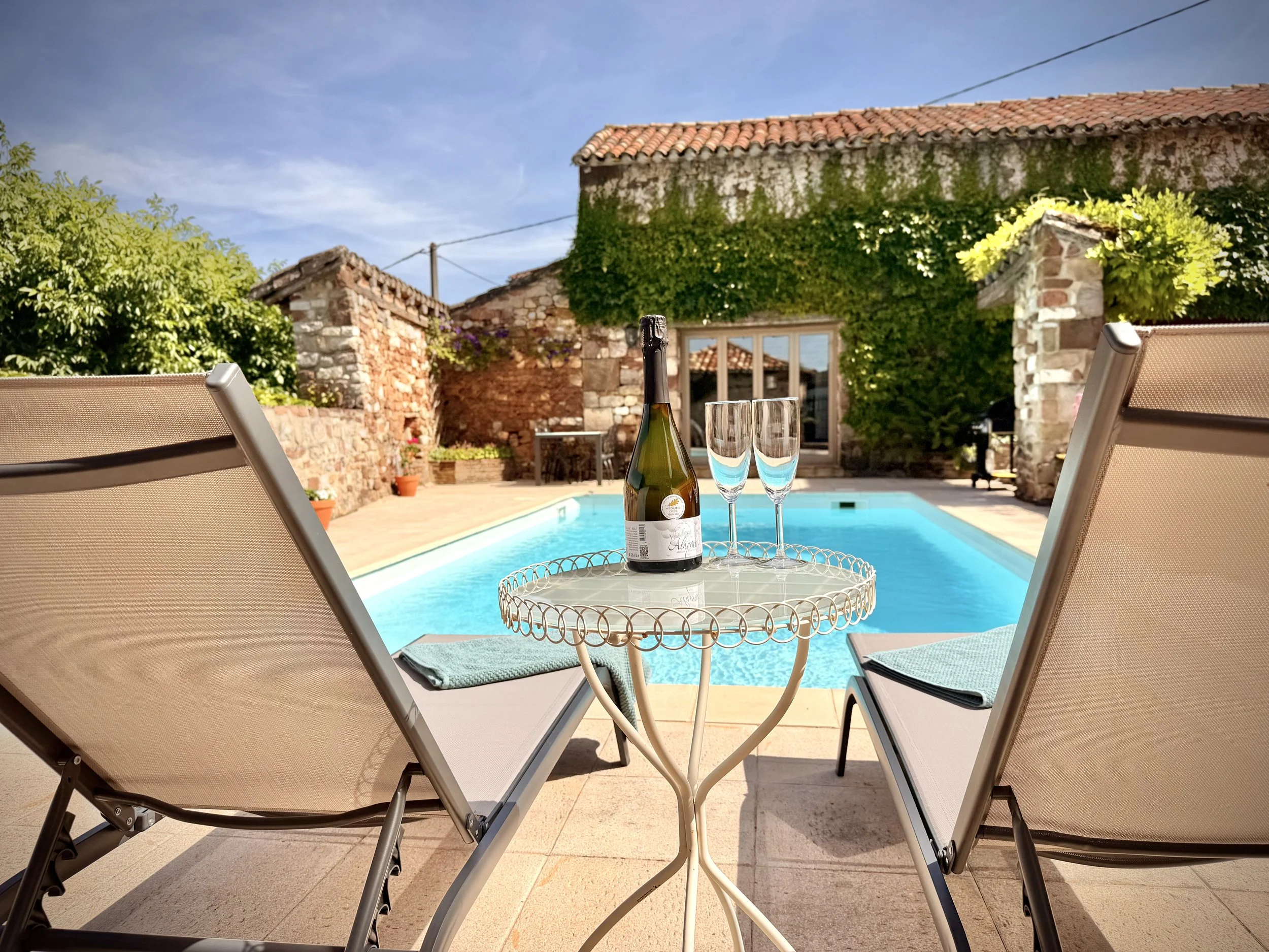 View of a backyard swimming pool with beige lounge chairs, a small round table with a bottle of champagne and two glasses, surrounded by stone walls, greenery, and a rustic house with tiled roof under a blue sky.
