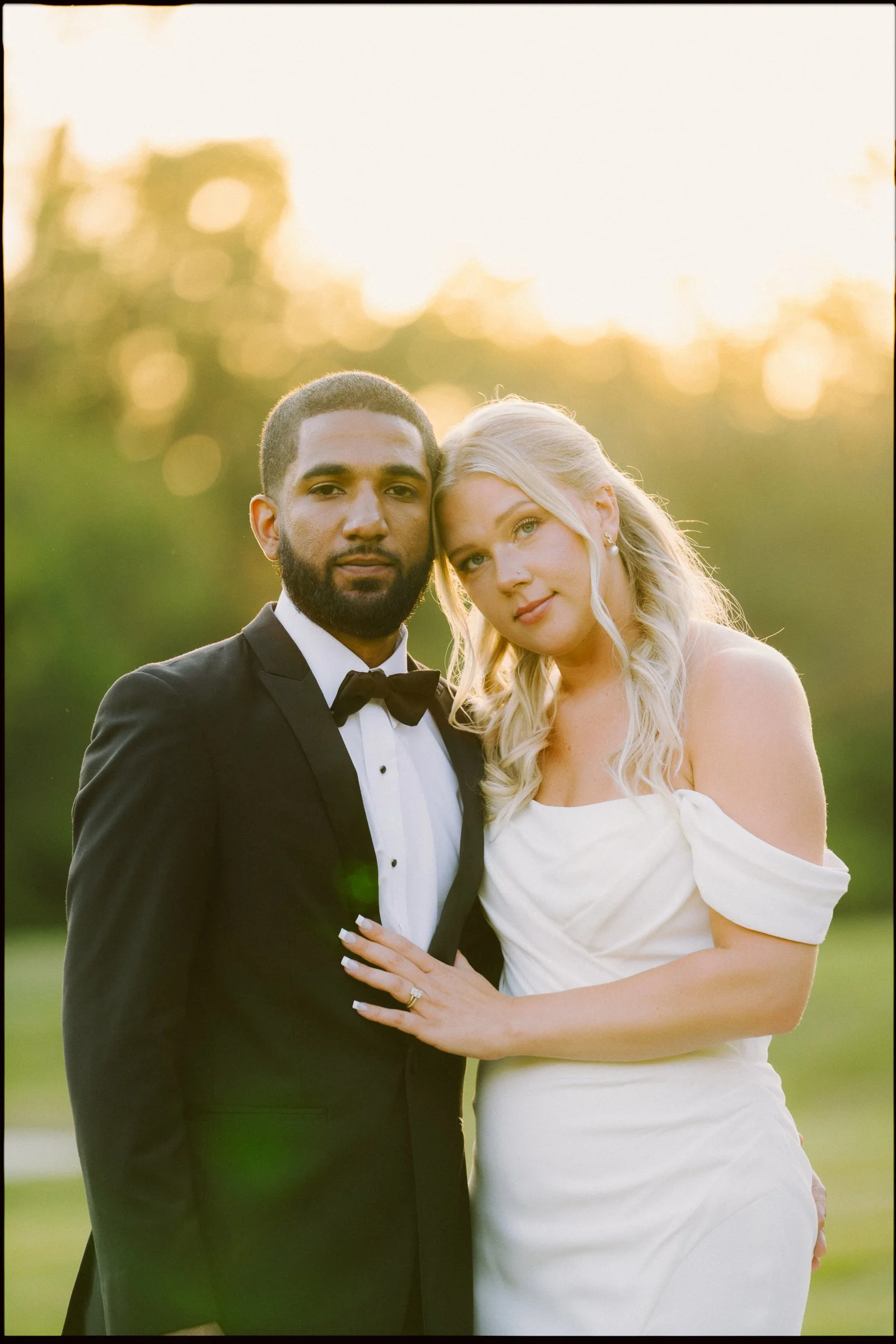 A bride and groom in wedding attire standing outdoors during sunset, with the bride resting her head on the groom's shoulder and both smiling softly.