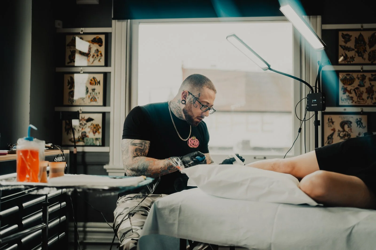 A tattoo artist with glasses and tattoos on his arms, working on a client in a tattoo studio. The studio has colorful artwork on the walls and a large window letting in natural light, with a bright overhead lamp providing additional lighting.