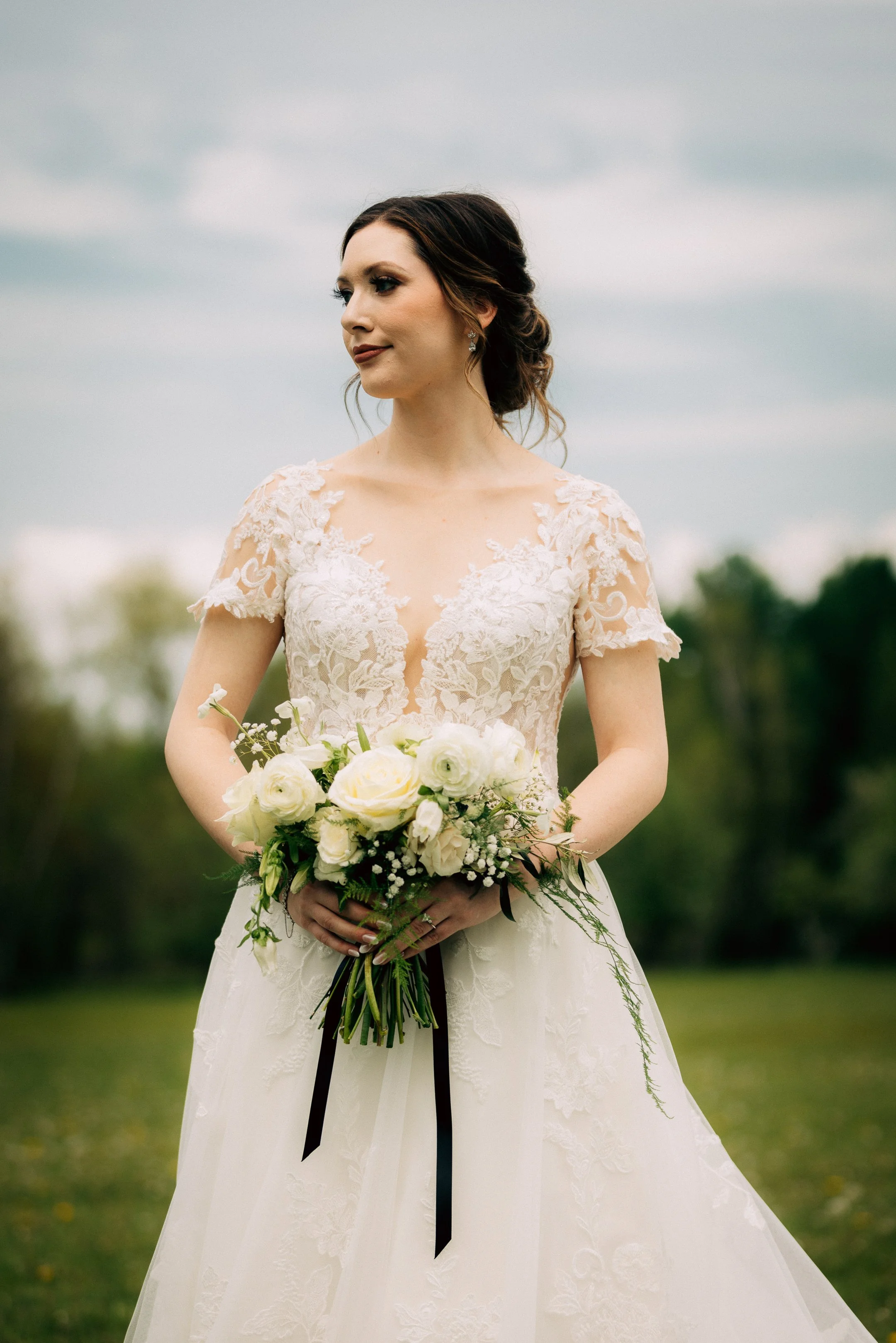 A bride with dark brown hair styled in an elegant updo, wearing a lace wedding gown with a deep neckline, holding a bouquet of white roses and greenery, outdoors on a cloudy day.