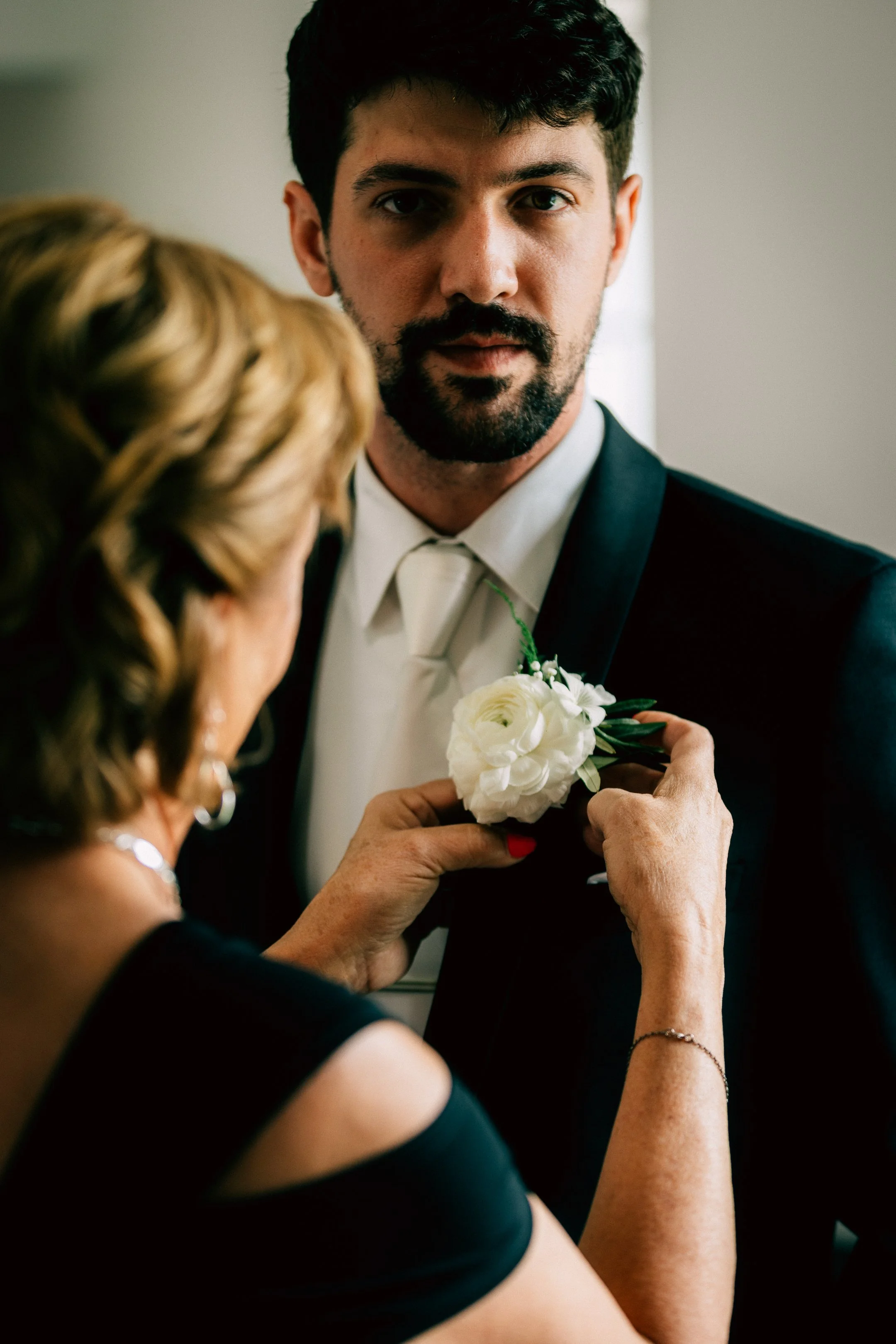 A man in a black tuxedo with a white shirt and a boutonniere is being helped by a woman with curly hair, possibly adjusting or pinning a white flower corsage on his tuxedo.