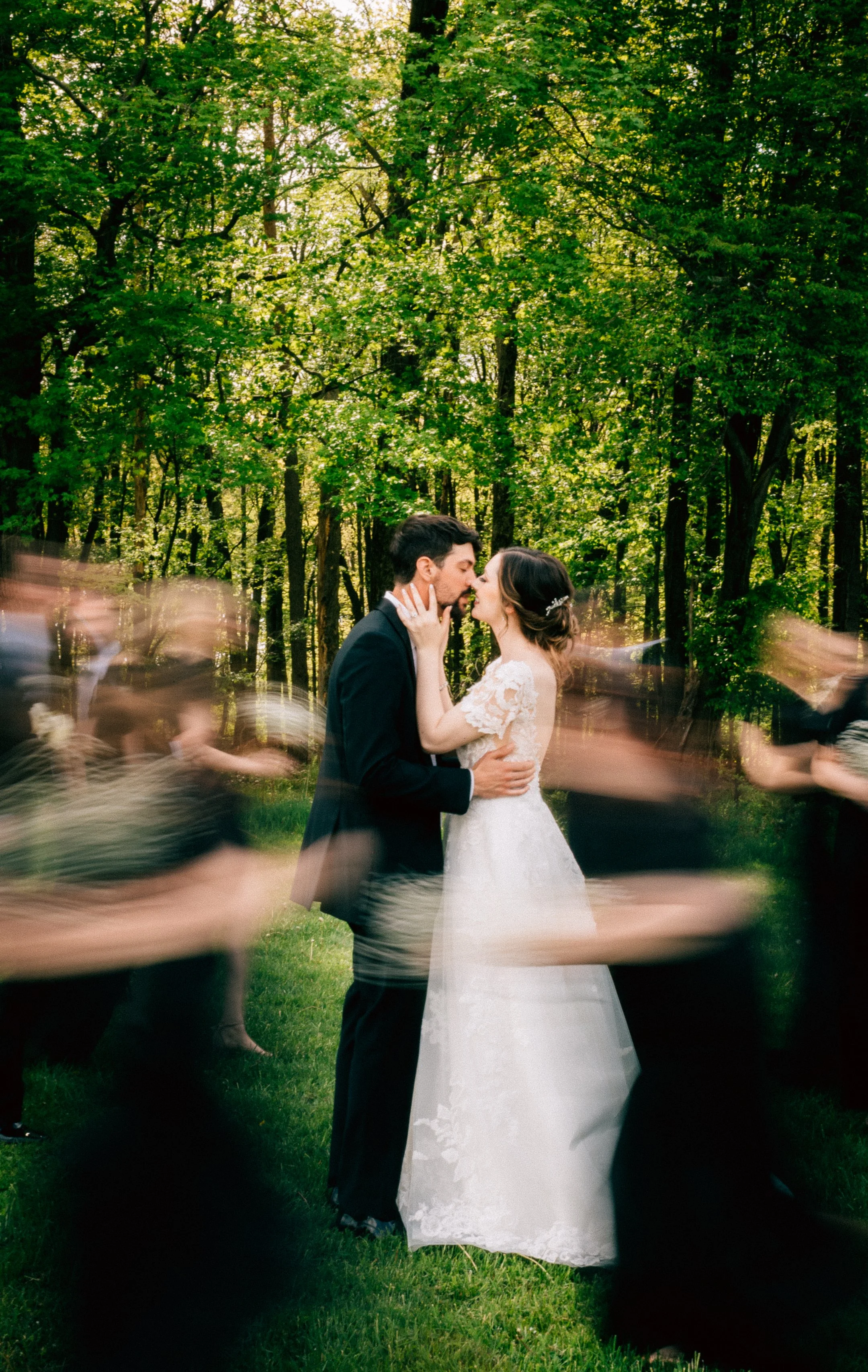 A bride and groom are kissing in a forested outdoor wedding setting with green foliage, surrounded by blurred guests in motion.