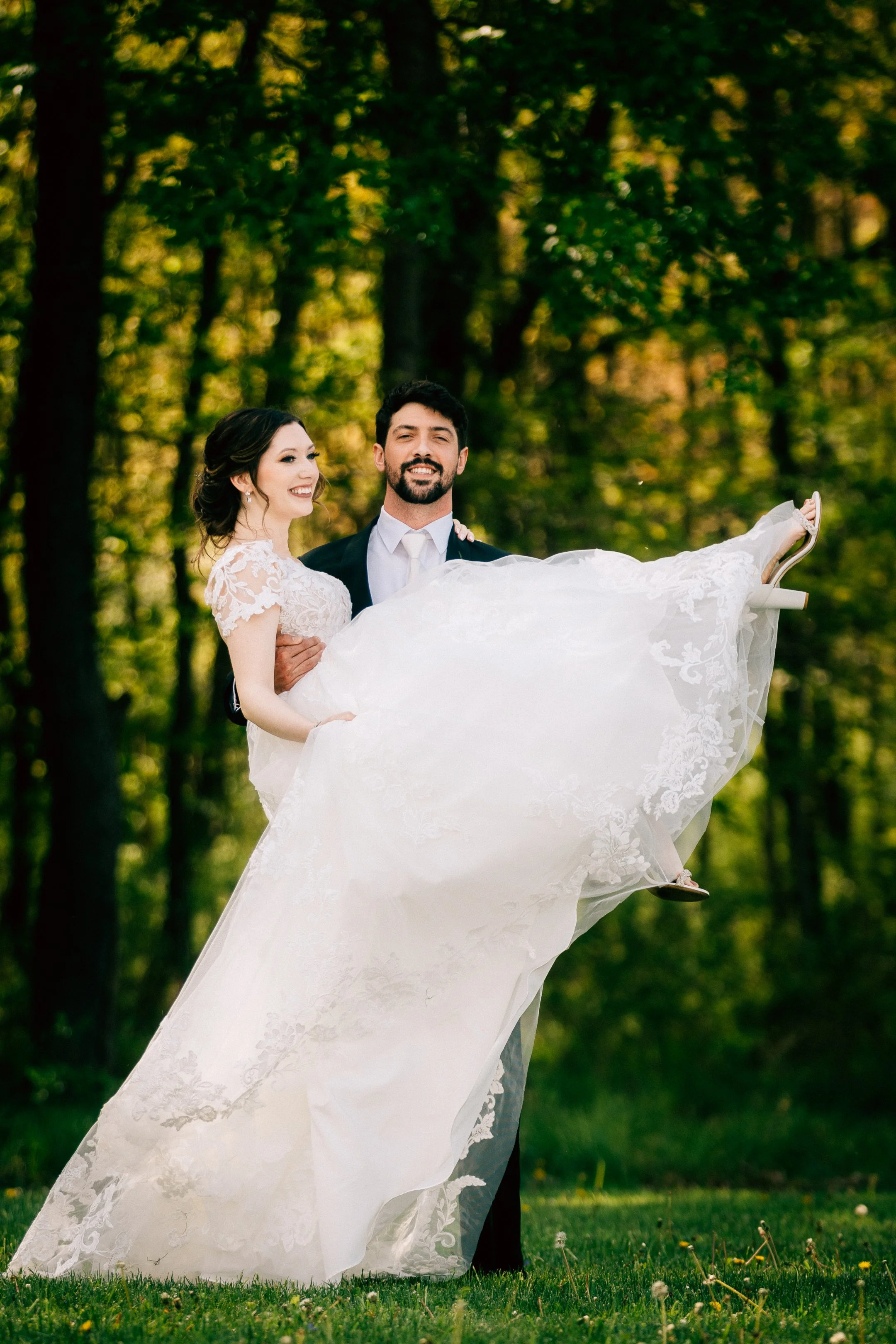 A couple dressed in wedding attire, with the groom holding the bride in his arms, standing outdoors on a grassy area with trees in the background.