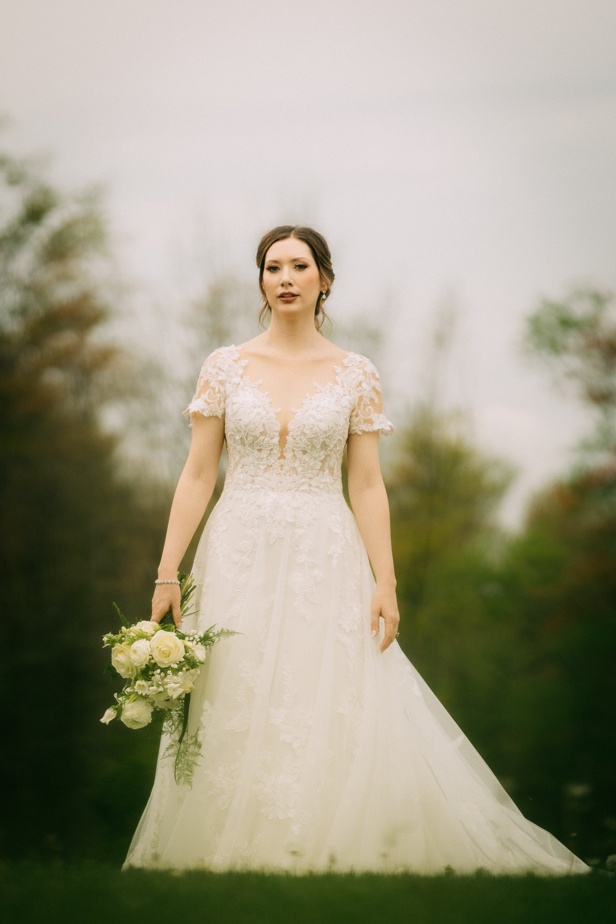 A bride in a white lace wedding dress holding a bouquet of white roses and greenery, standing outdoors with blurred trees in the background.