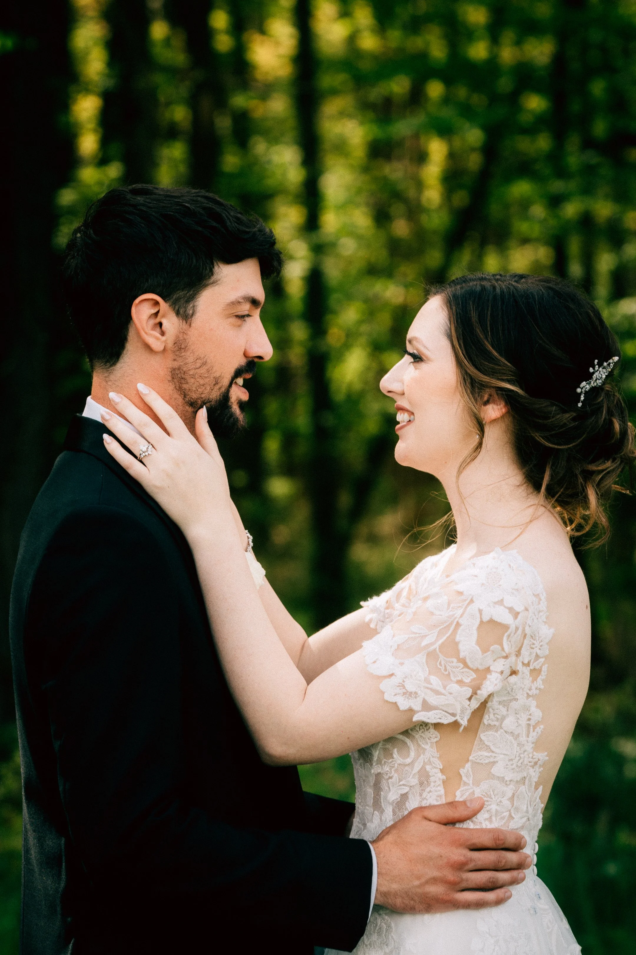 A bride and groom holding each other closely outdoors in a forest, smiling at each other, with the bride's hand on the groom's face showing an engagement ring.
