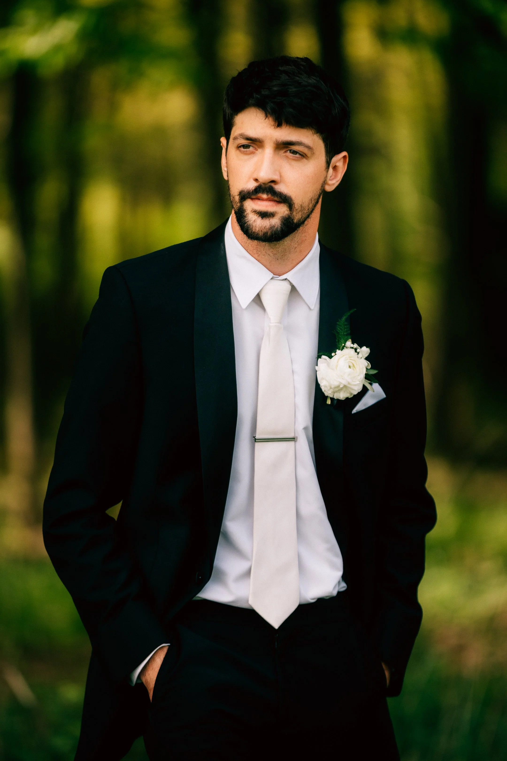 A man in a black tuxedo with a white shirt, tie, and boutonnière stands outdoors in a forested area with blurred green trees in the background.