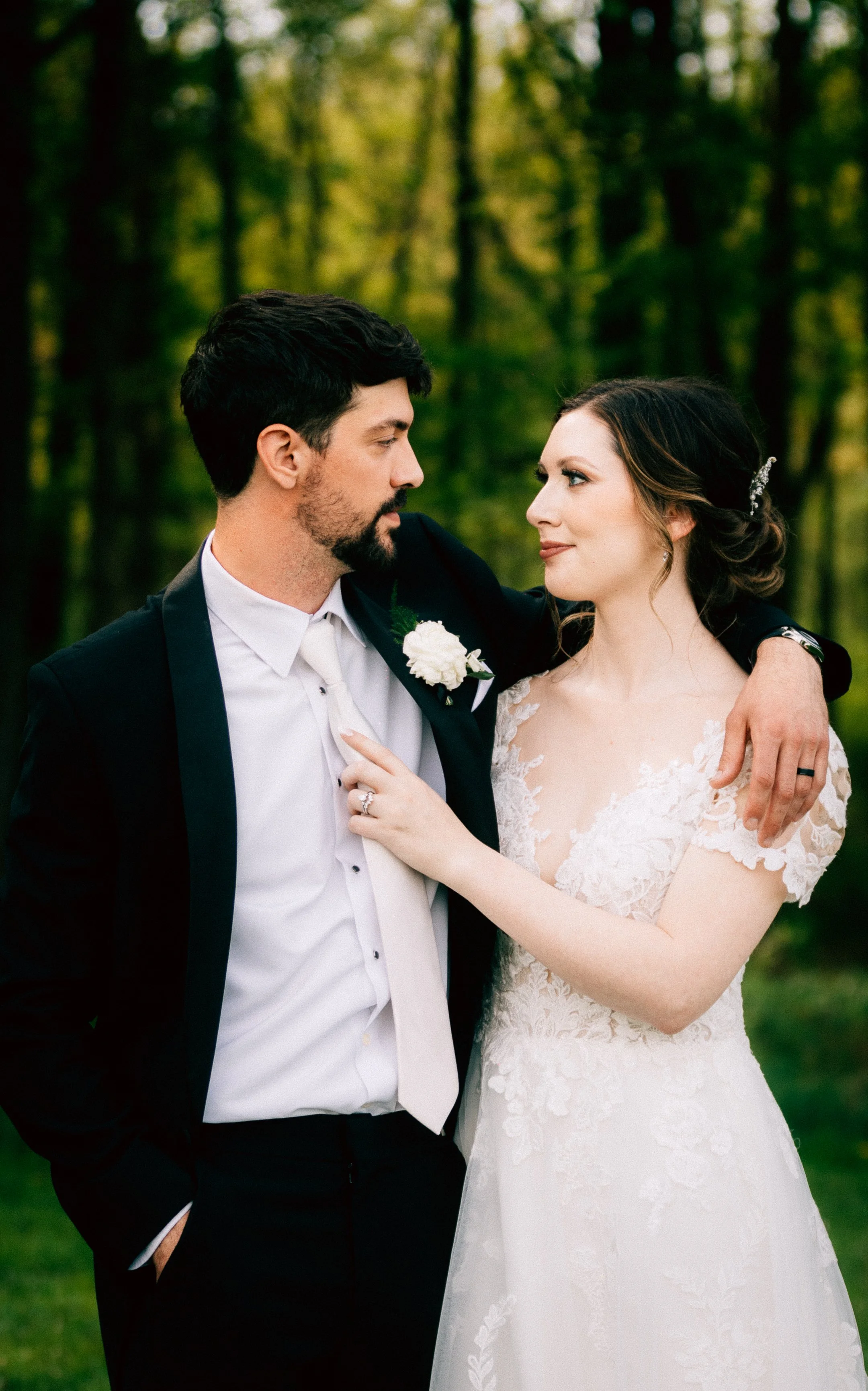 A bride and groom in wedding attire standing outdoors in a wooded area, looking into each other's eyes.