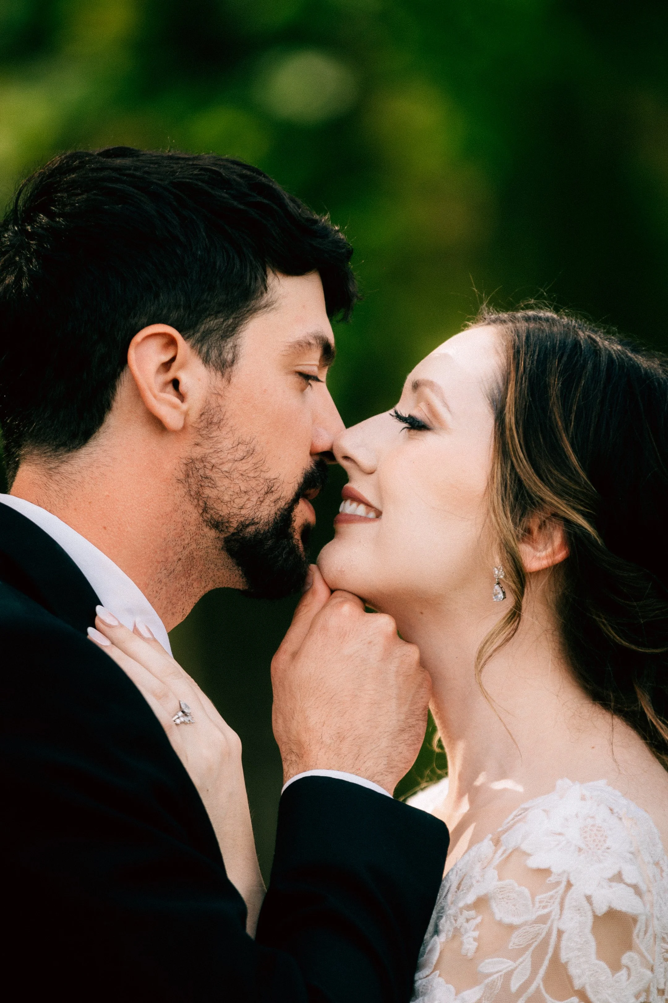 A bride and groom share an intimate moment outdoors, with eyes closed and foreheads touching. The groom, with dark hair and beard, wears a black tuxedo; the bride wears a white lace wedding dress and earrings.