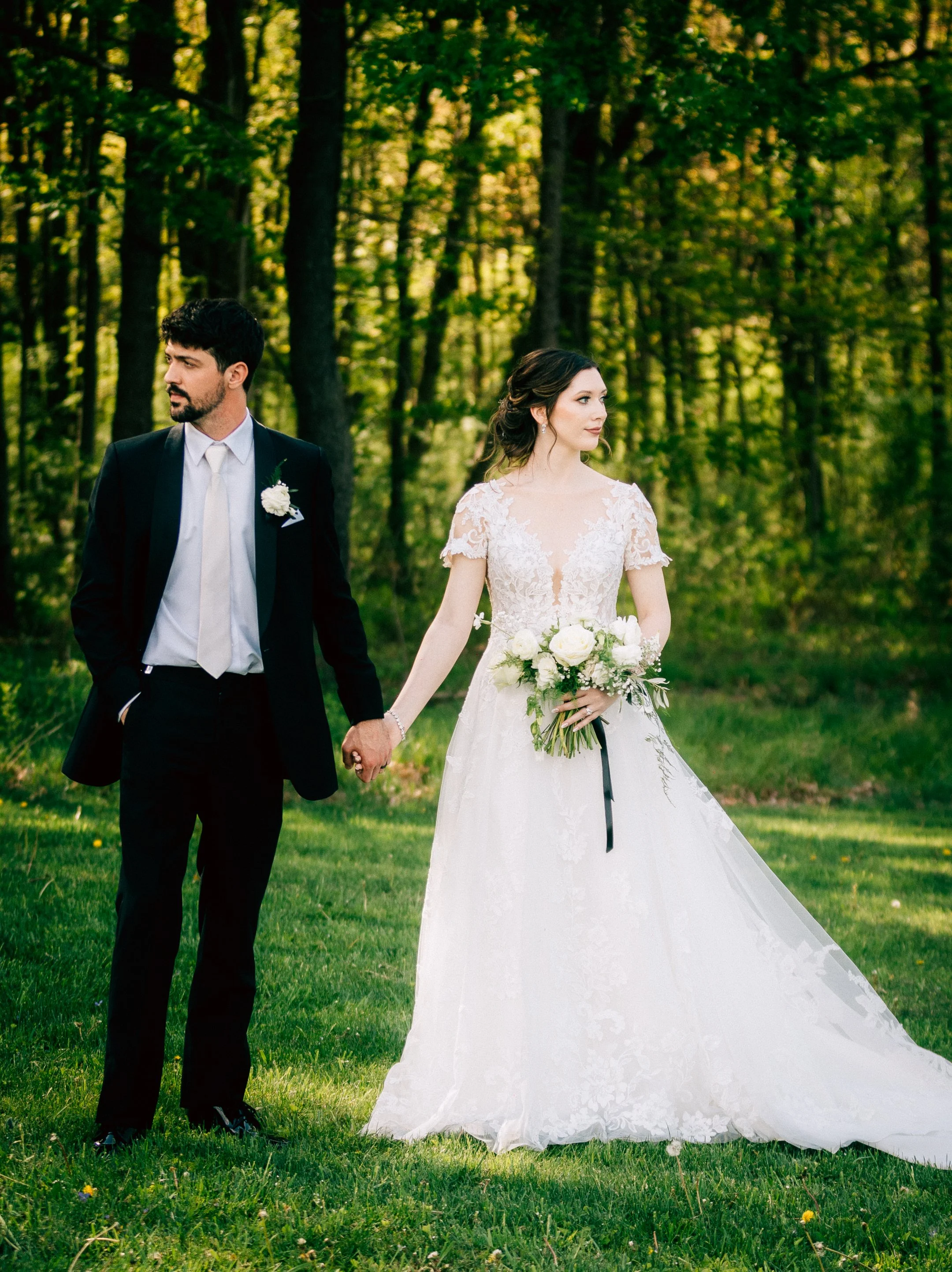 Bride in a white wedding dress holding a bouquet and groom in a black suit holding hands in a wooded outdoor setting.