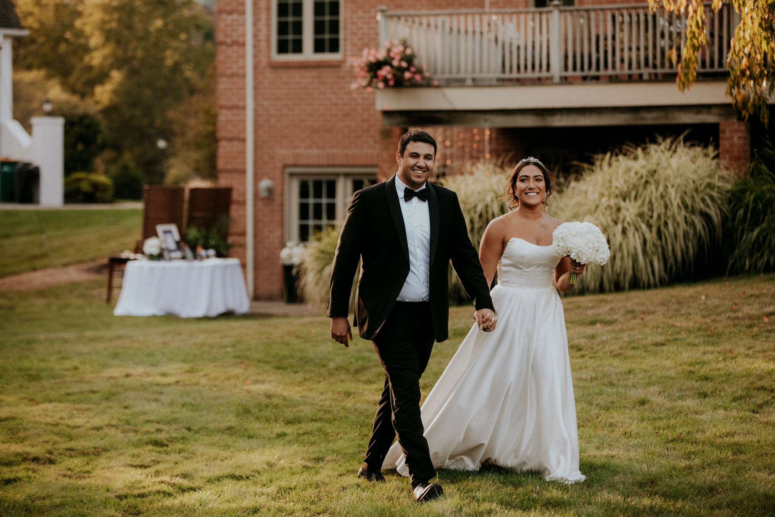 A bride and groom walking hand-in-hand outdoors during a wedding celebration. The bride is wearing a strapless white wedding gown and holding a bouquet of white flowers, while the groom is dressed in a black tuxedo with a bow tie. They are smiling an