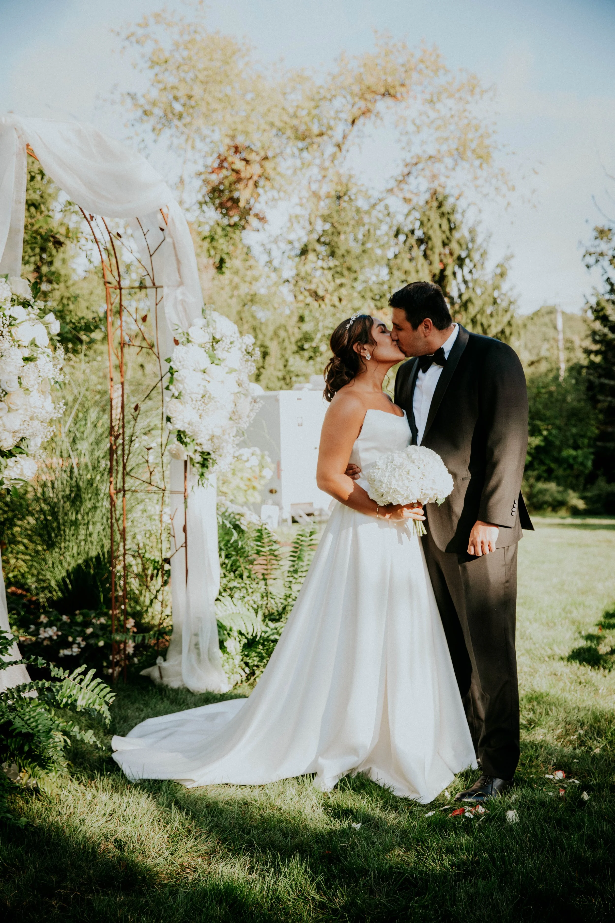 A bride and groom sharing a kiss outdoor during their wedding ceremony, with floral arrangements and greenery surrounding them.