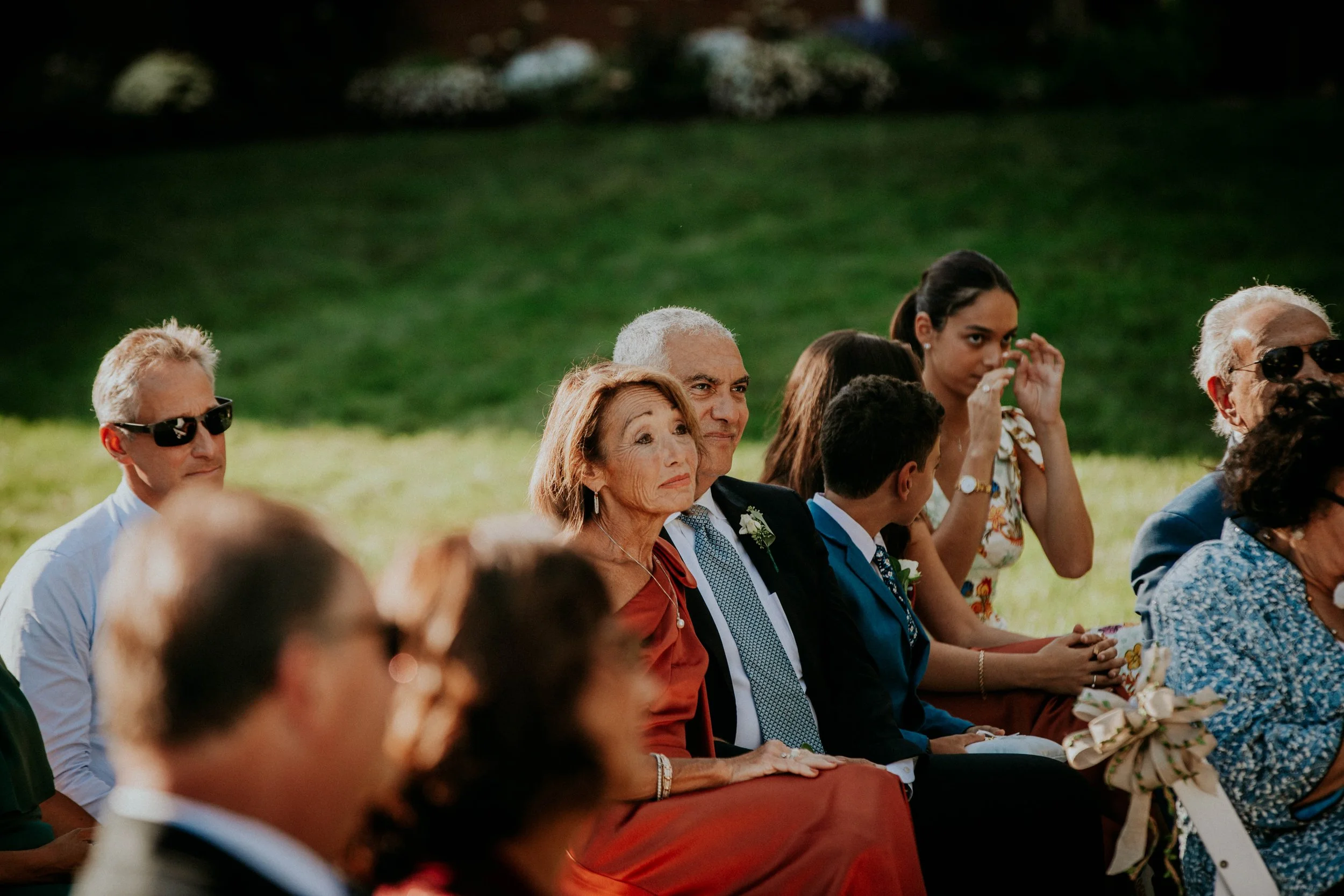 A group of people dressed in formal attire sitting outdoors on a sunny day, possibly at a wedding or event, with greenery and trees in the background.