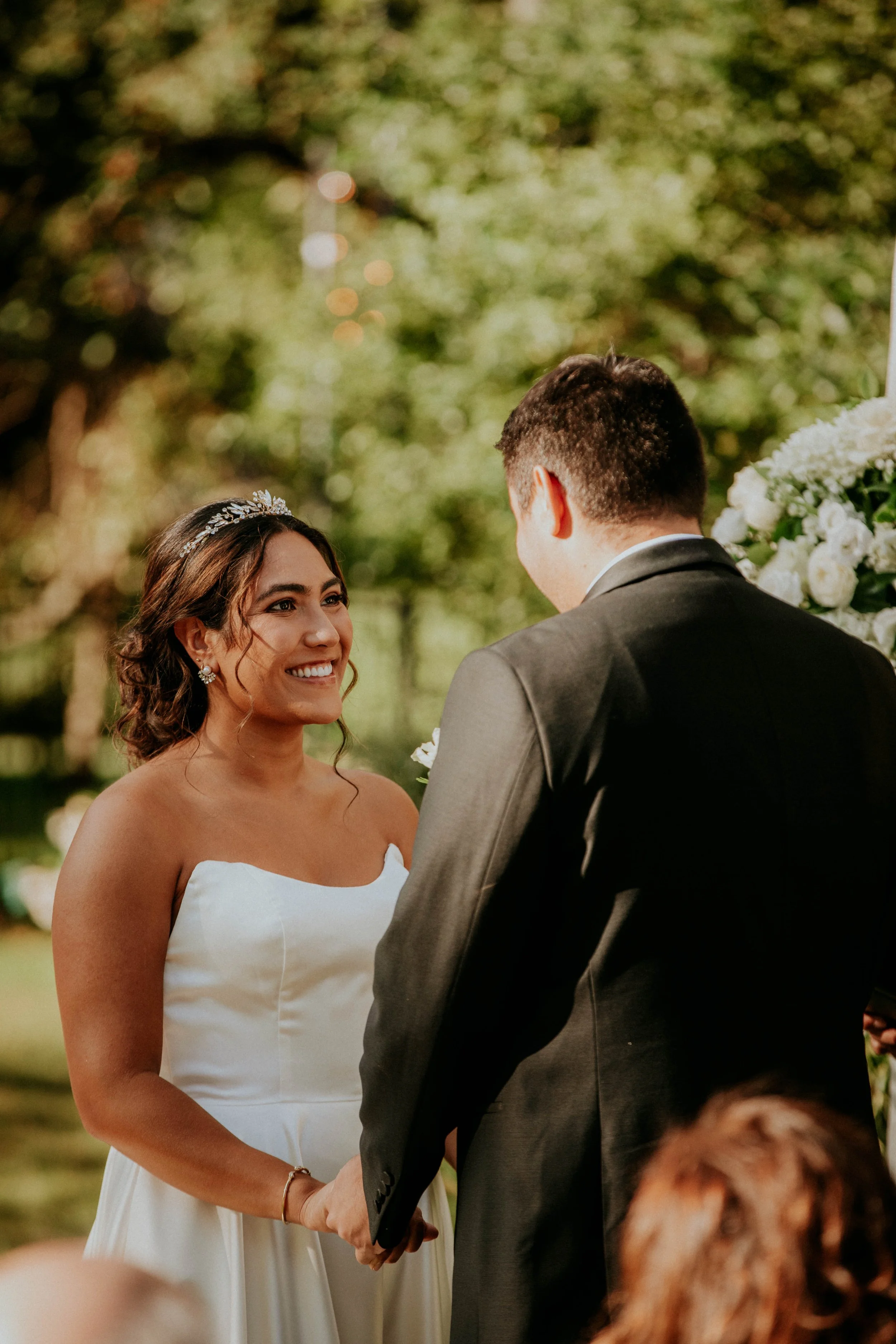 A bride and groom during their wedding ceremony, holding hands and gazing at each other, outdoors with lush greenery in the background.