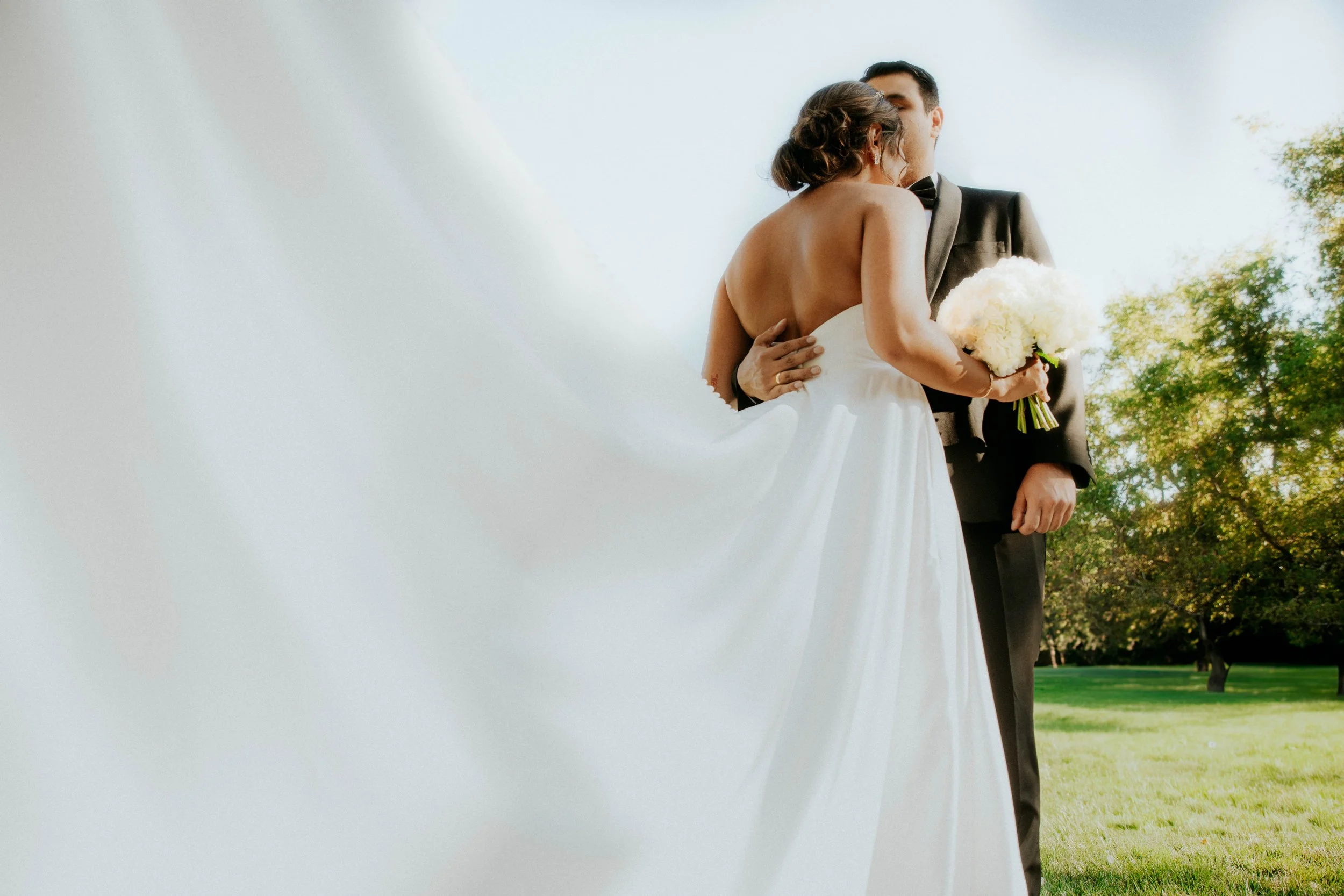A bride and groom embracing outdoors during a wedding, with the bride holding a bouquet of white flowers.