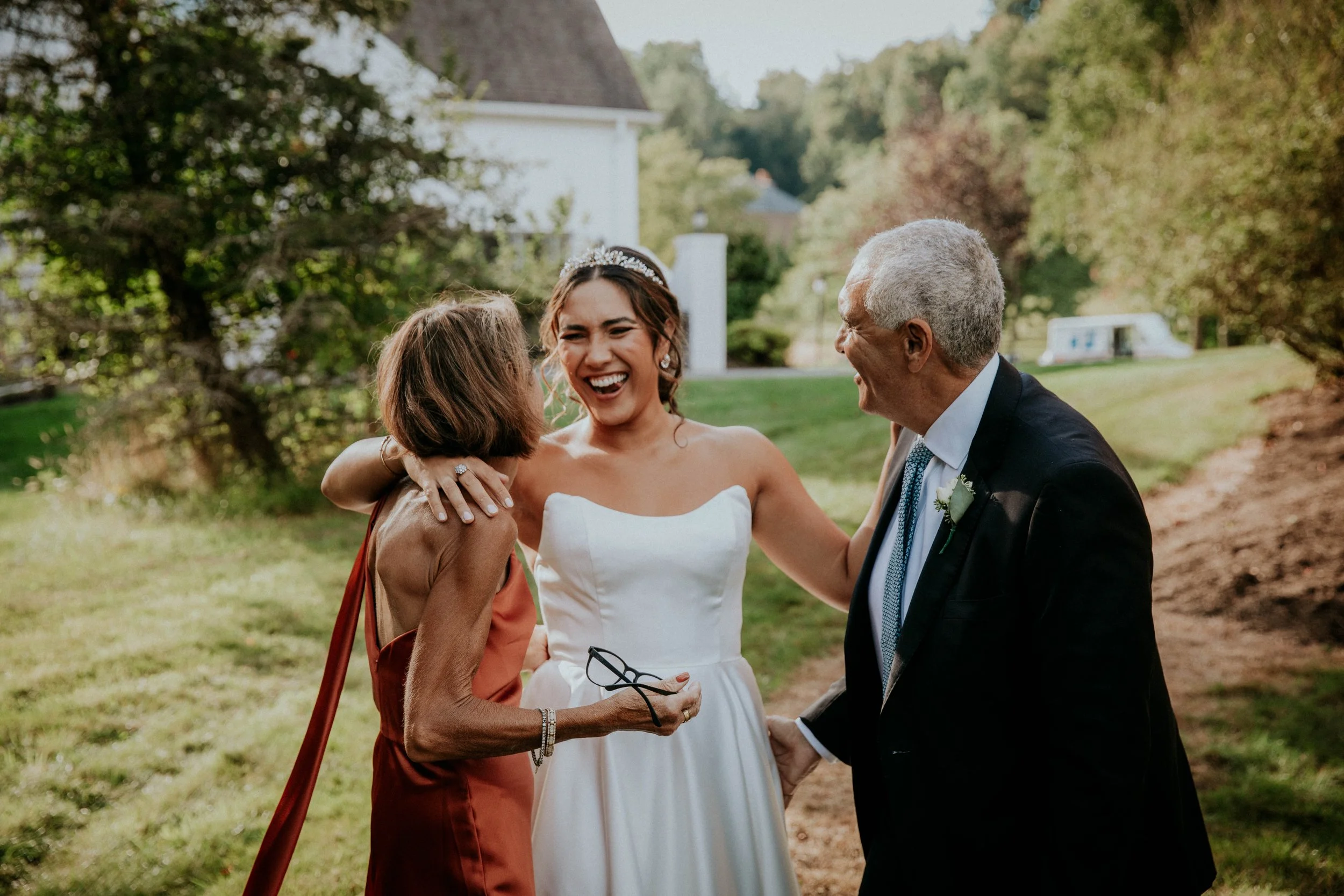 A bride in a white wedding dress smiling and hugging an older woman in a rust-colored dress, while an older man in a suit stands nearby and smiles, outdoors in a lush green setting.