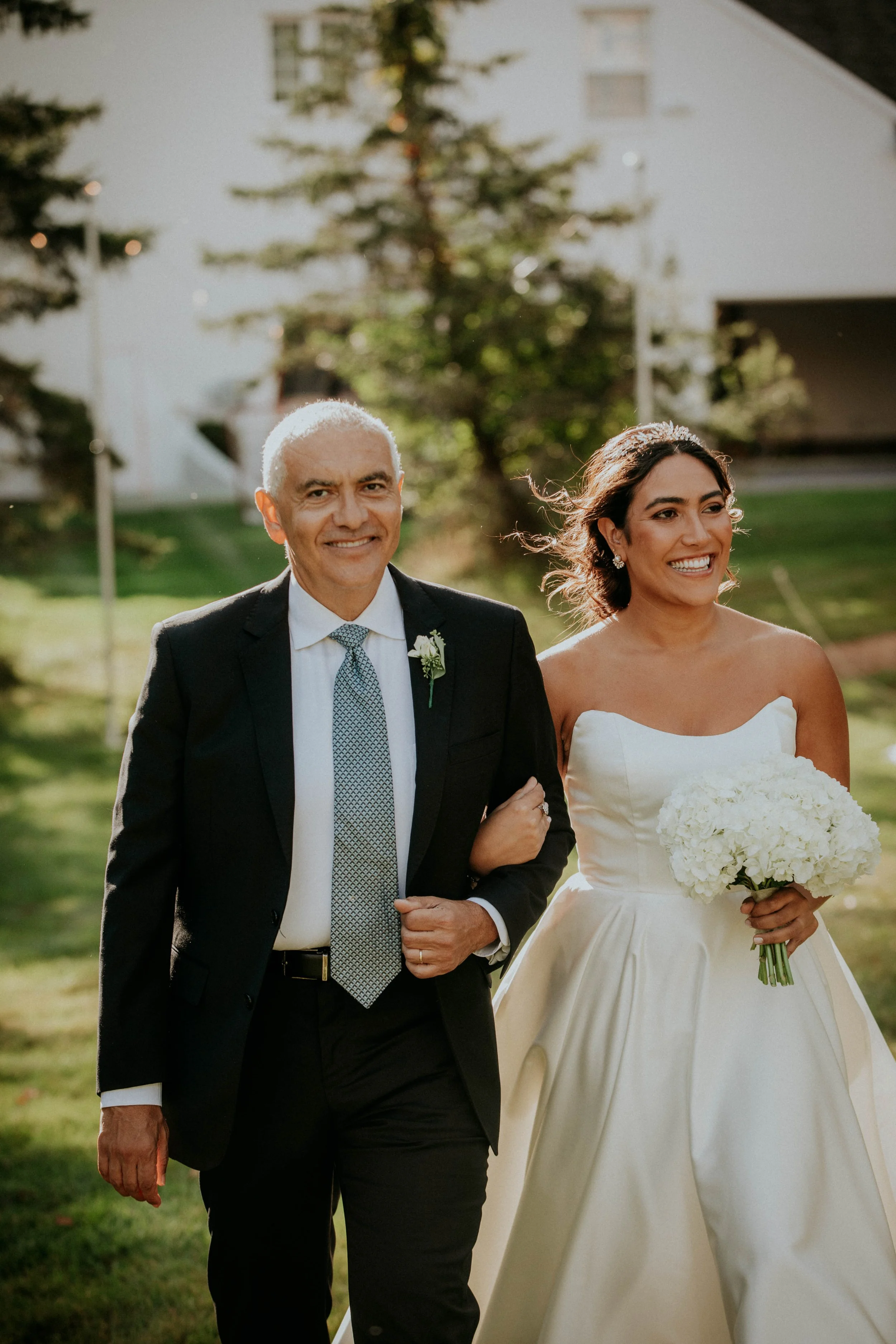 A smiling older man in a black suit and a young woman in a white wedding dress walking outdoors in a park-like setting with trees and buildings in the background.