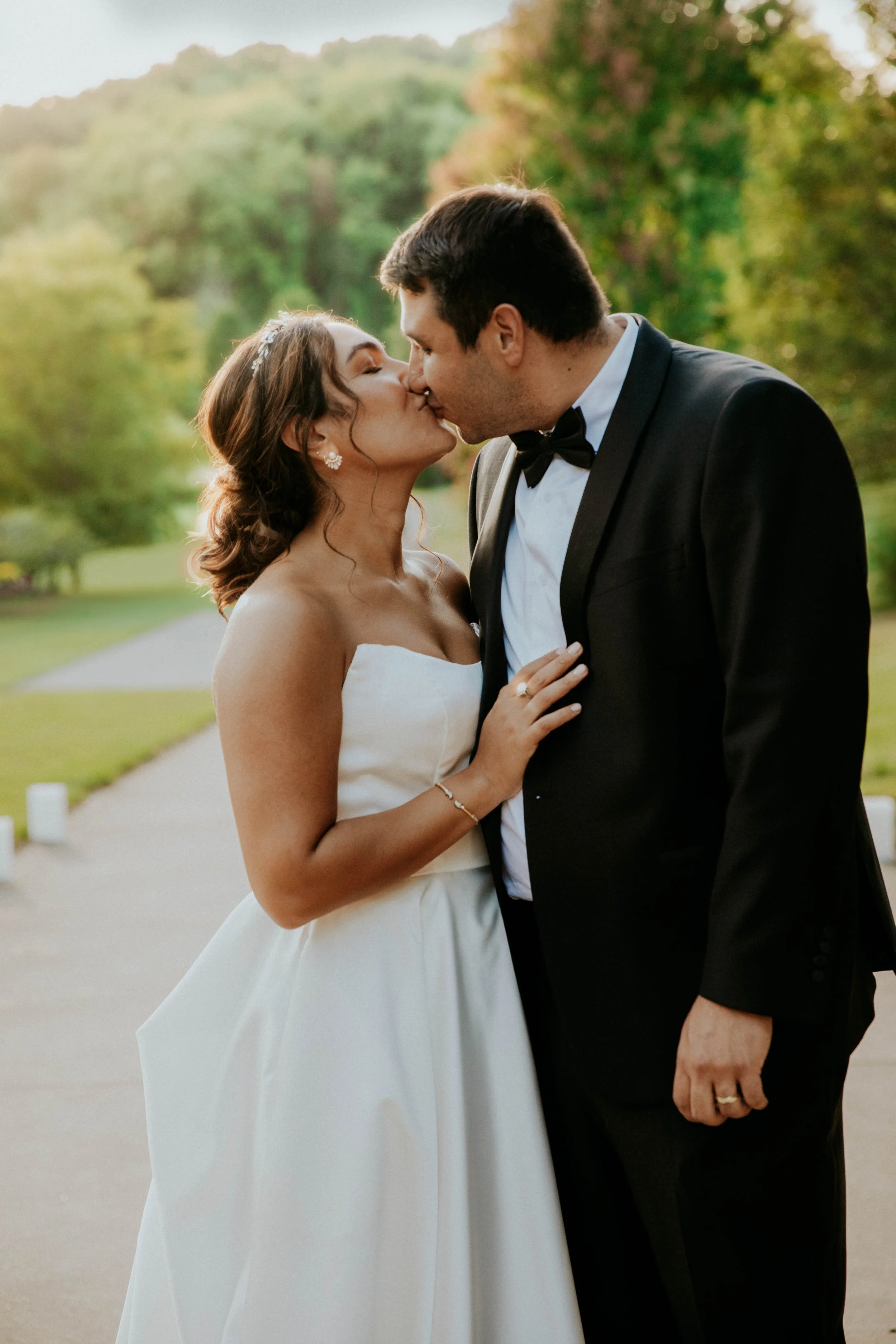 A bride and groom sharing a kiss outdoors during daytime, with the bride in a strapless white gown and the groom in a black tuxedo with a bow tie, surrounded by green trees.