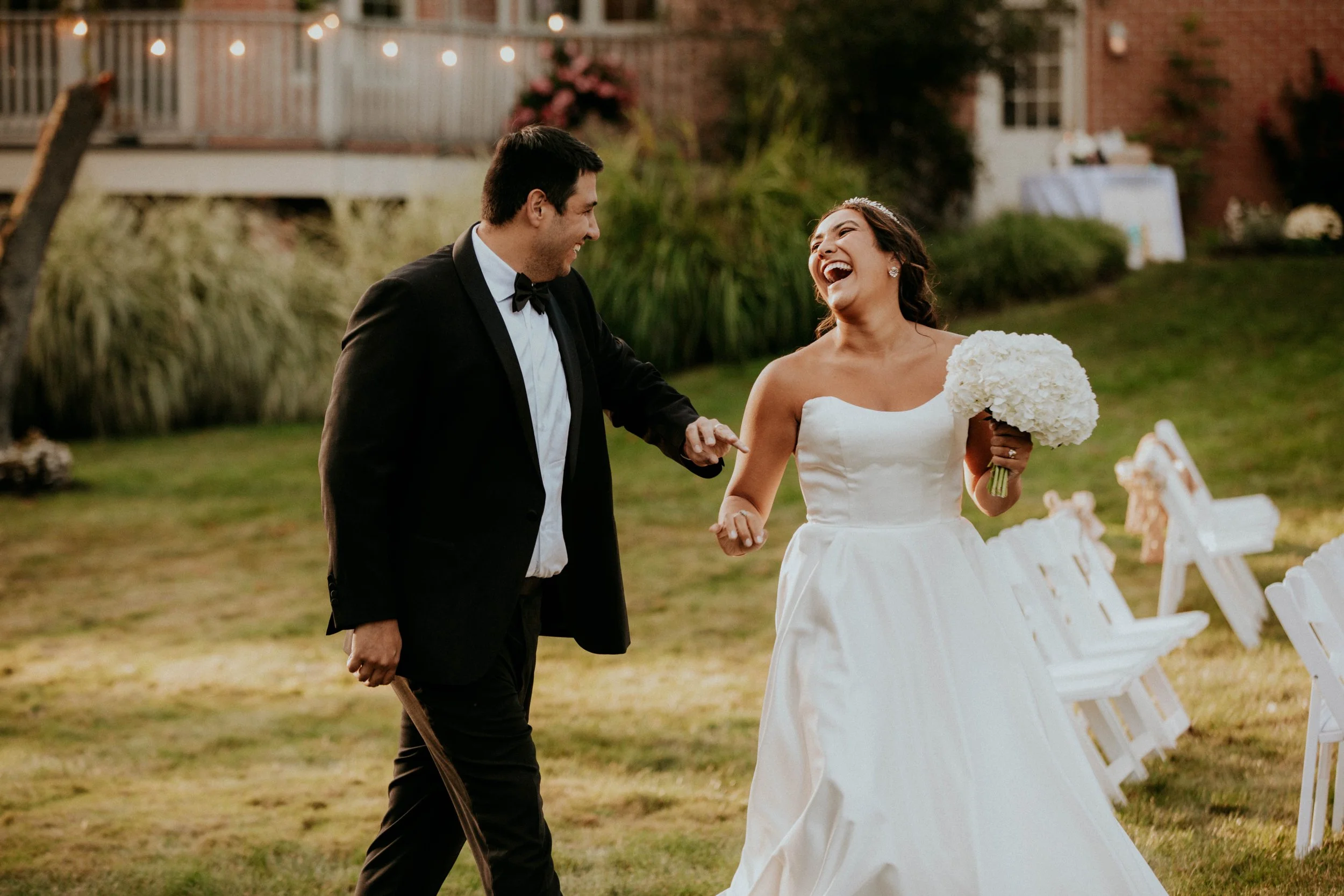 A joyful bride and groom holding hands and laughing during their outdoor wedding celebration in the evening, with wedding decor visible in the background.