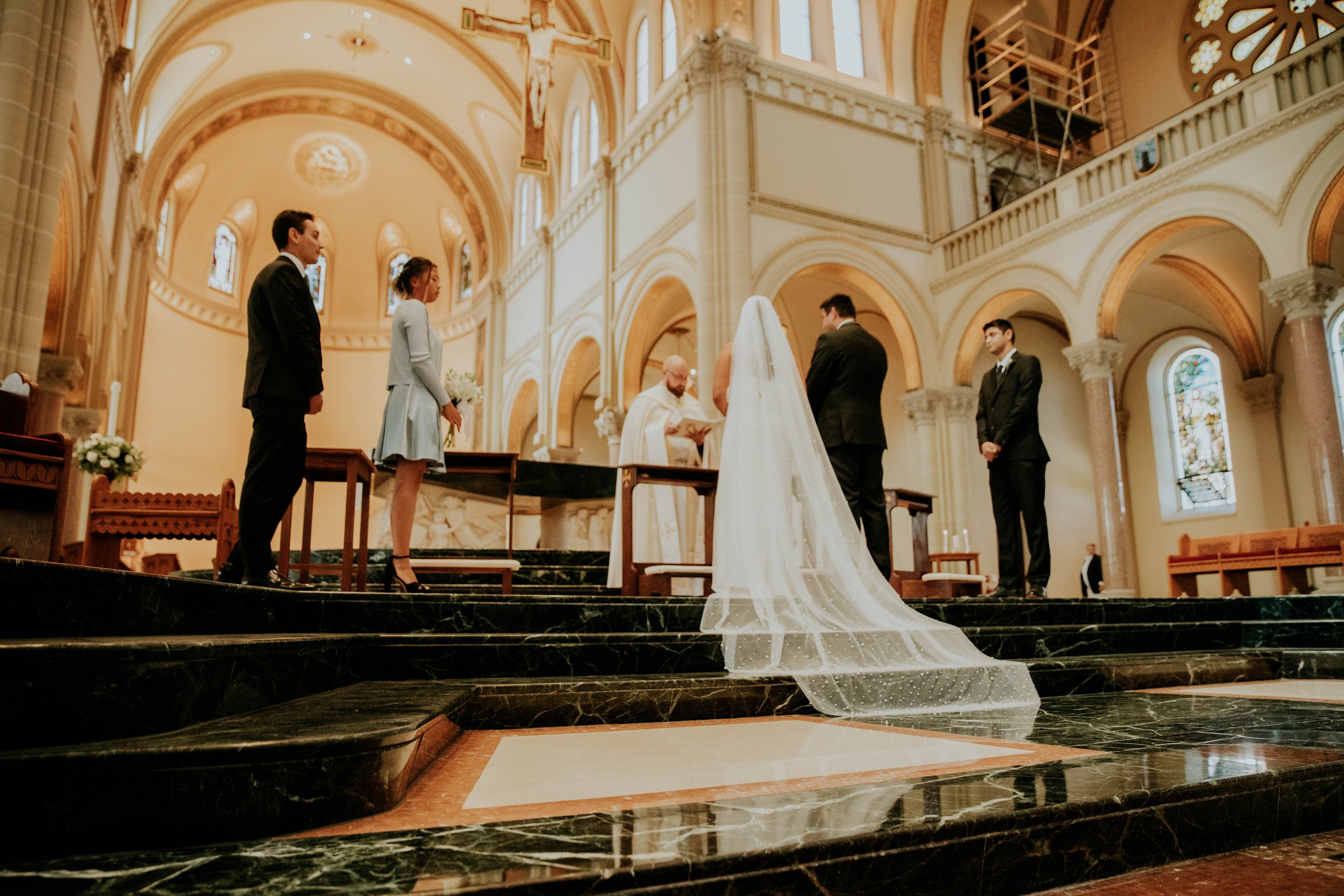 A wedding ceremony inside a church with a bride and groom standing before an officiant, surrounded by bridesmaids and groomsmen, with stained glass windows and ornate architecture in the background.