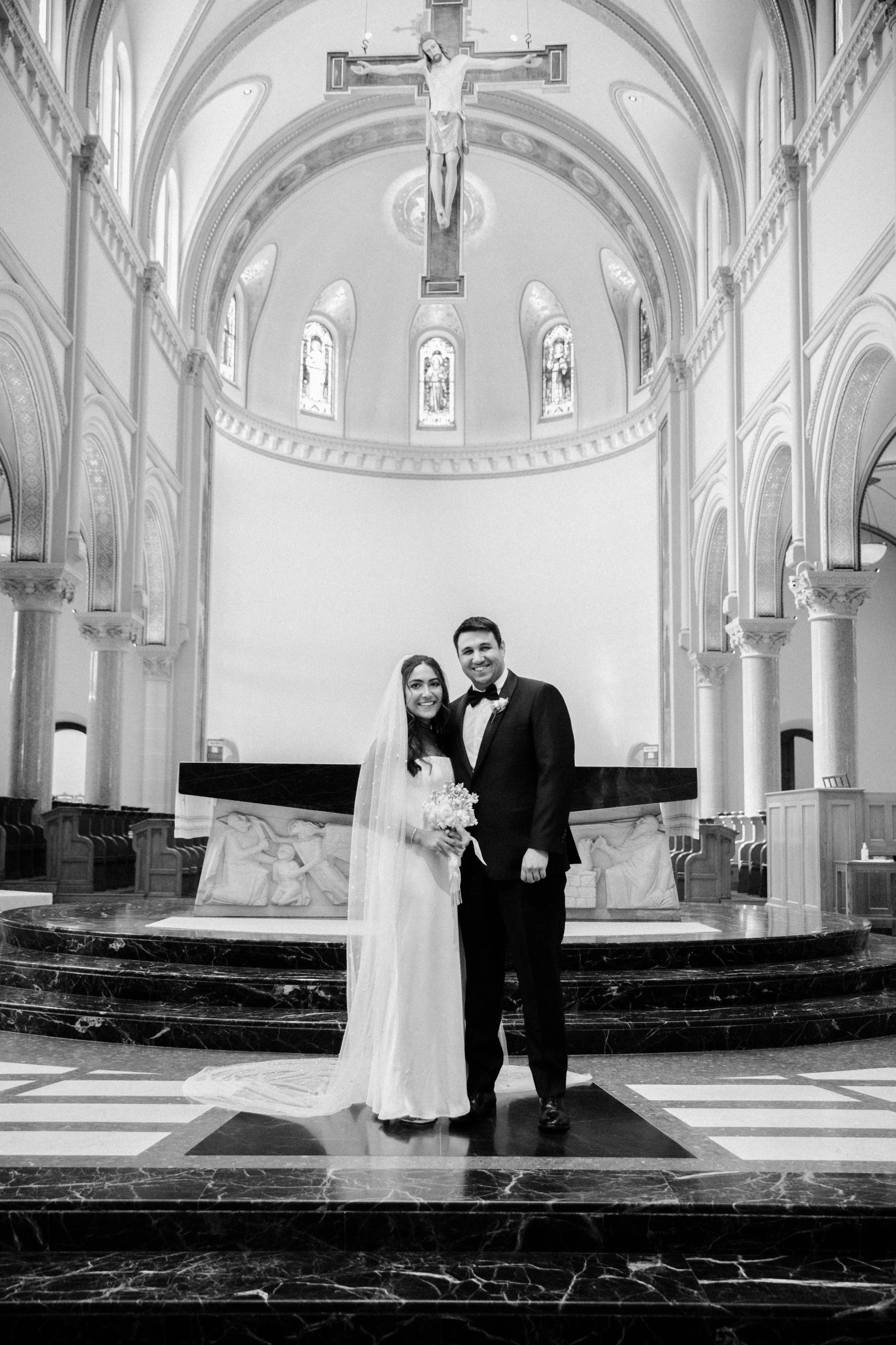 A bride and groom standing together inside a church, smiling for a wedding photo. The bride is wearing a wedding dress and holding a bouquet, while the groom is wearing a tuxedo. The church has high ceilings, arched windows, and a crucifix hanging ab