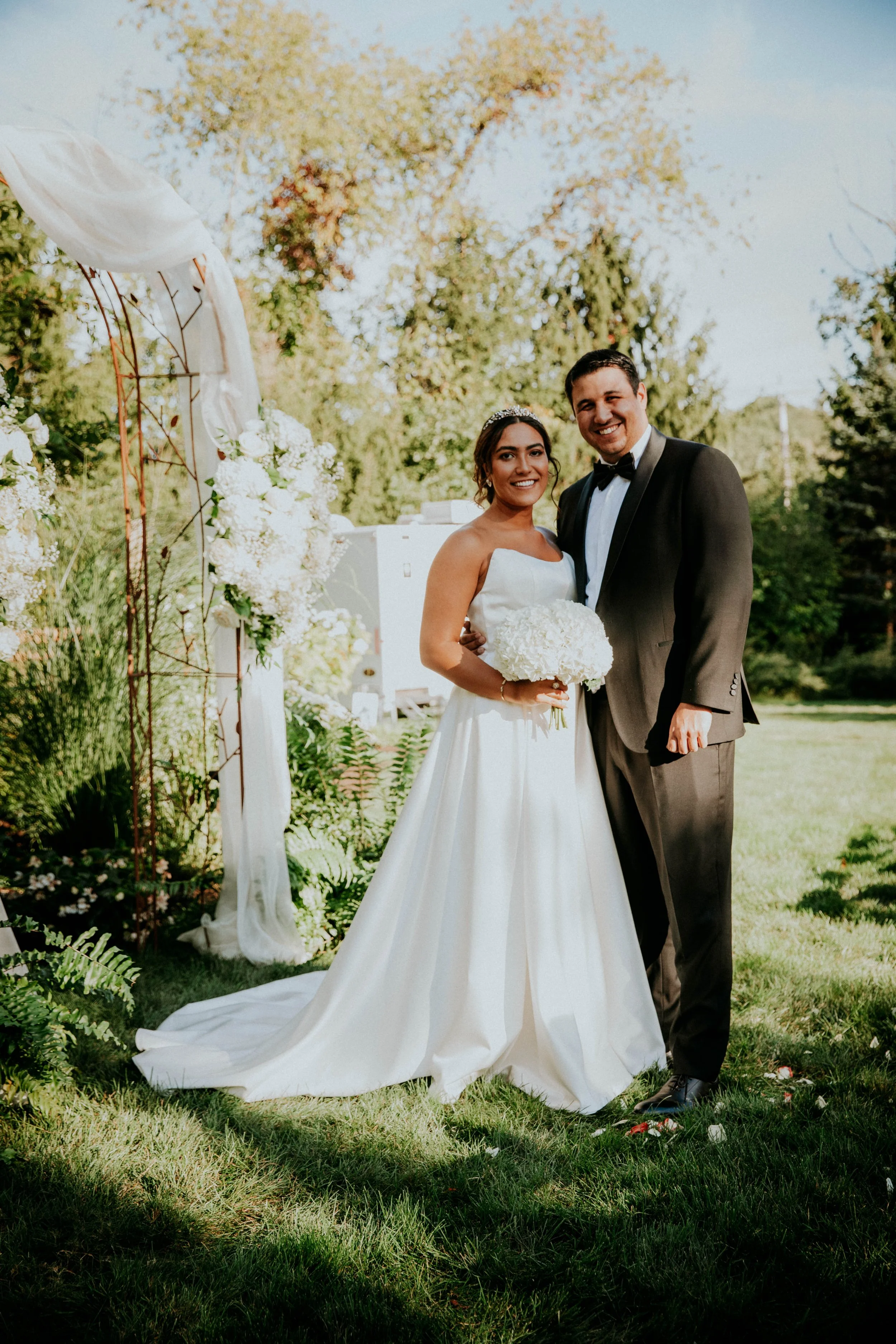A bride and groom standing outdoors on a wedding day, smiling at the camera. The bride is holding a bouquet of white flowers, wearing a white wedding dress with a train, and a tiara. The groom is in a black tuxedo with a bow tie. They are standing in