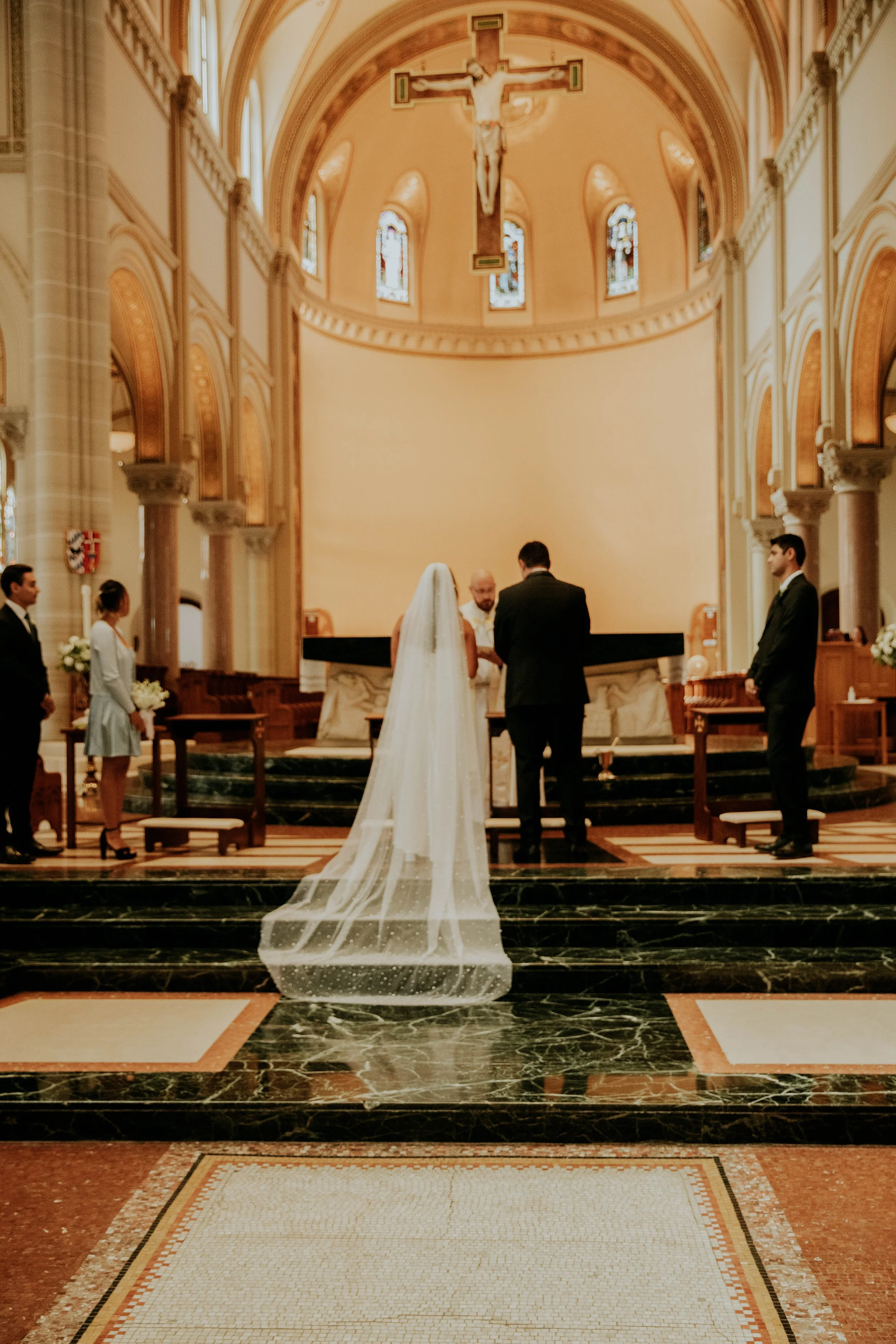 A wedding altar inside a church with a bride and groom standing before the officiant, surrounded by wedding attendants. The church features high arches, stained glass windows, and a large crucifix hanging above the altar.