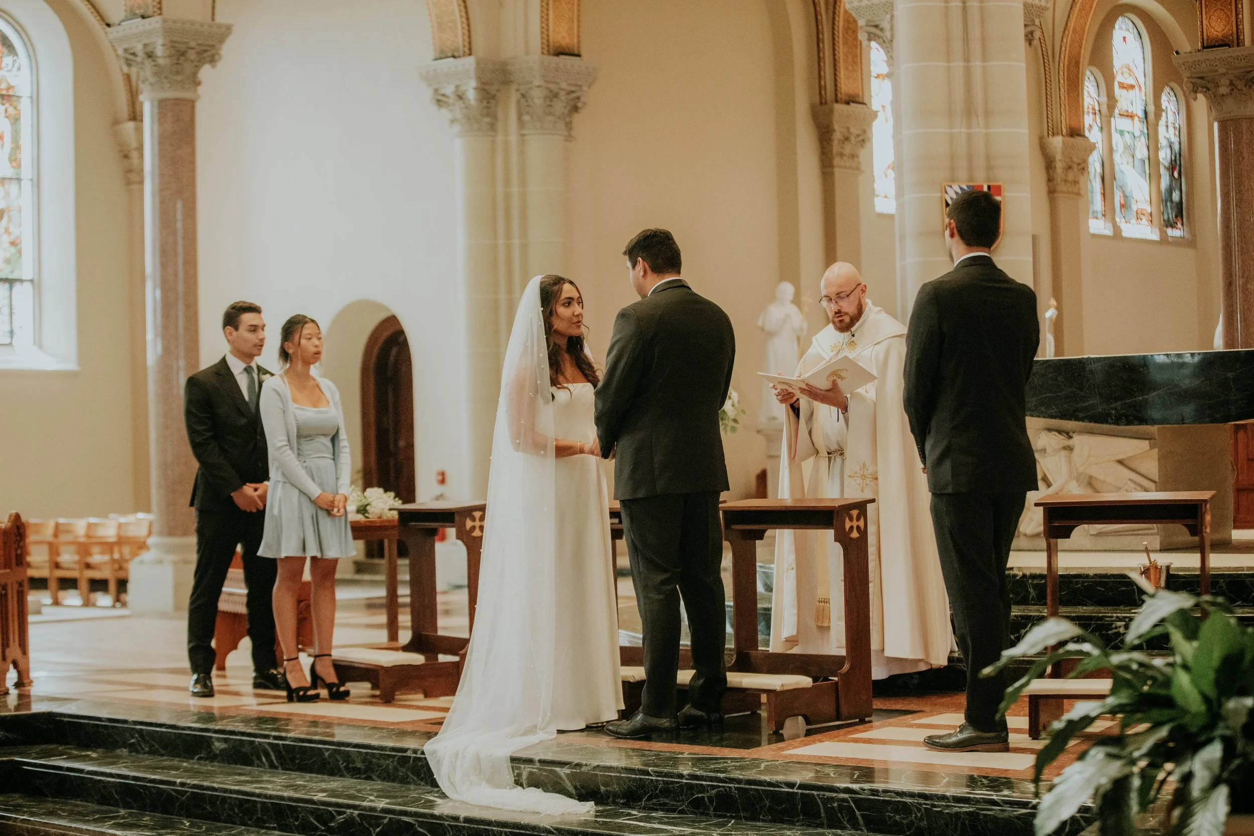 A couple exchanging vows in a church during a wedding ceremony, with a priest overseeing the event and three attendants standing nearby.
