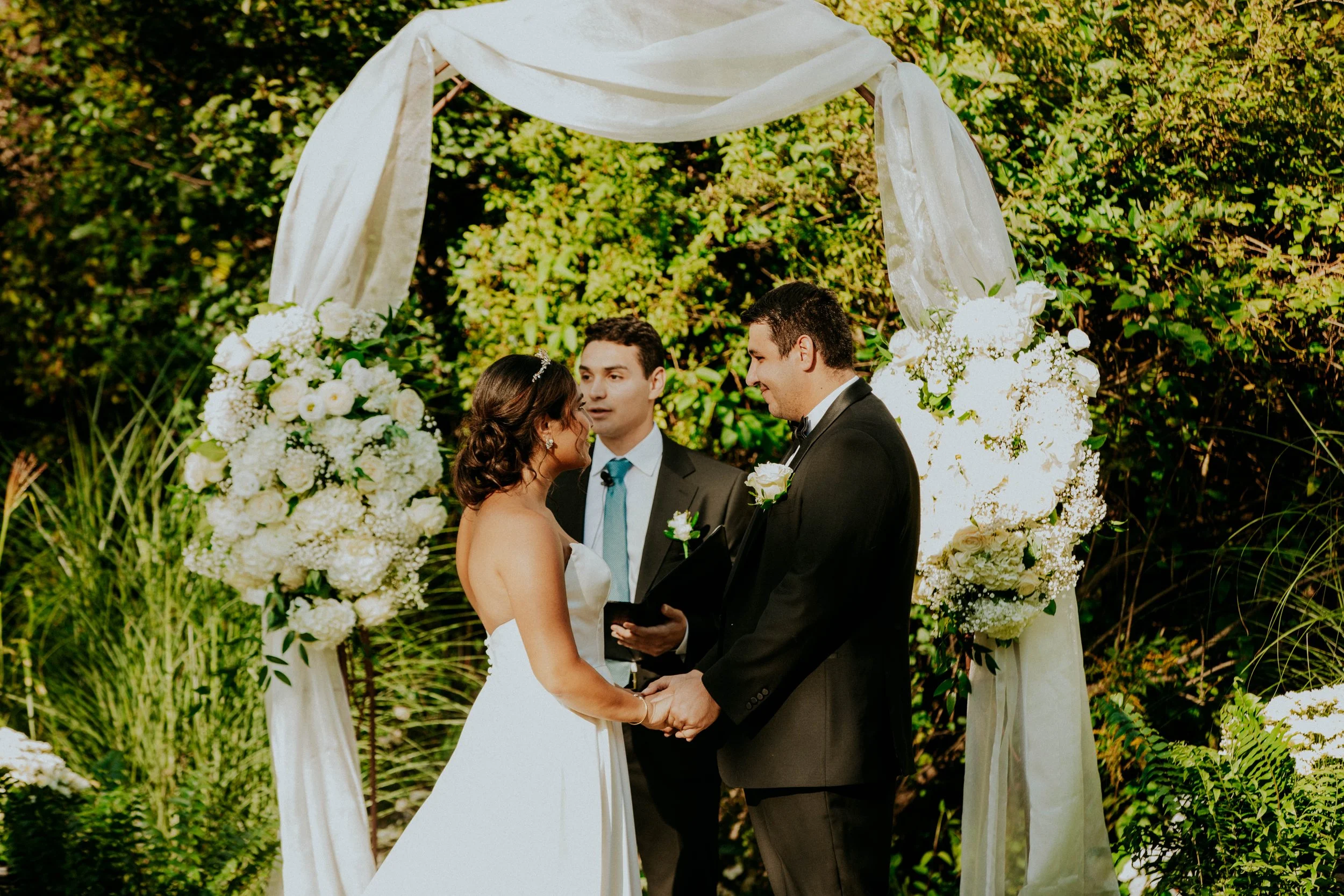 A bride and groom at their wedding ceremony, standing face to face, holding hands, under a white fabric arch decorated with white flowers, with a lush green foliage background.