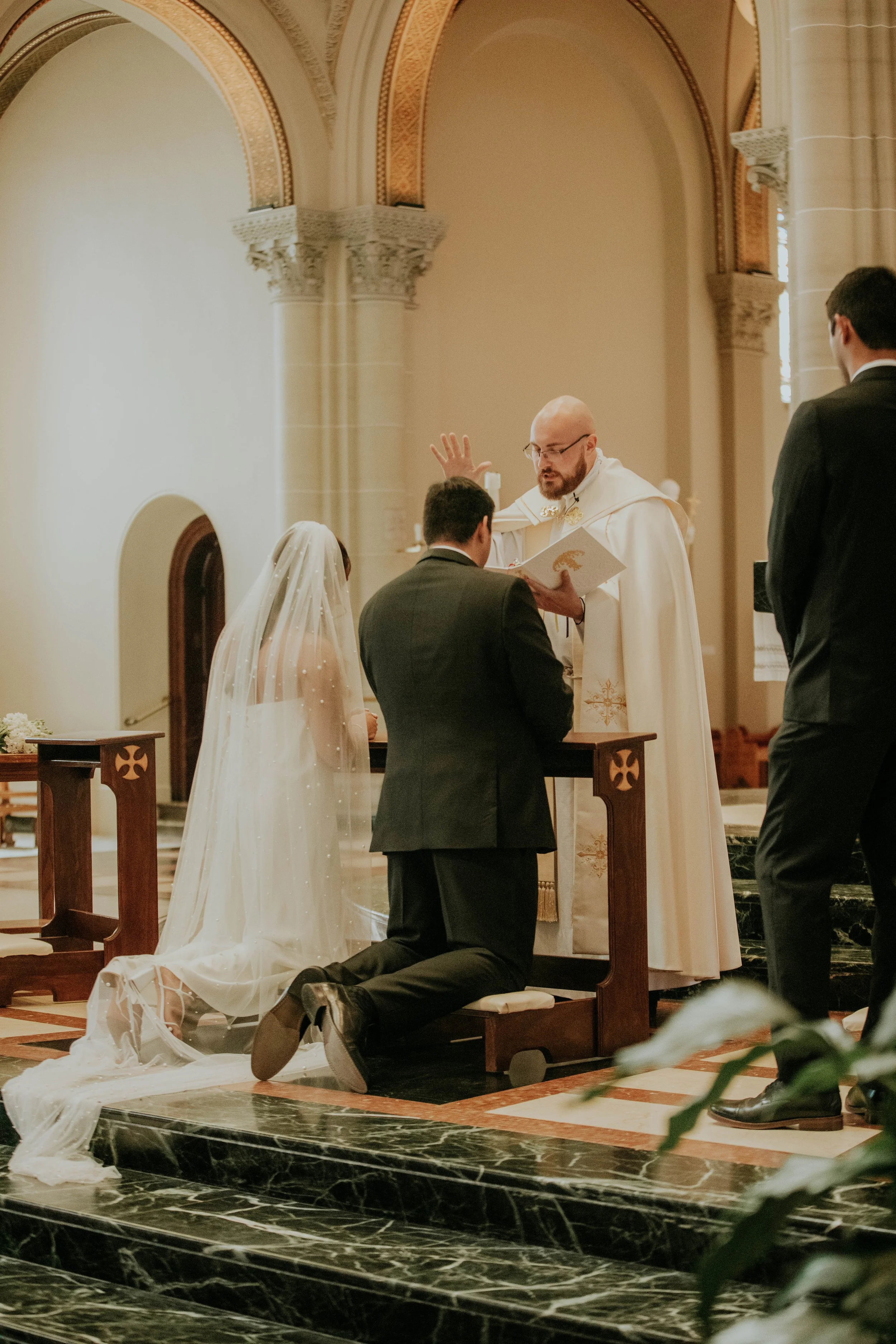 A wedding ceremony inside a church with a priest conducting the vows in front of a couple, a woman in a white wedding dress with a veil and a man in a black suit. Another man in a black suit is standing nearby, and a person kneels on the steps in fro