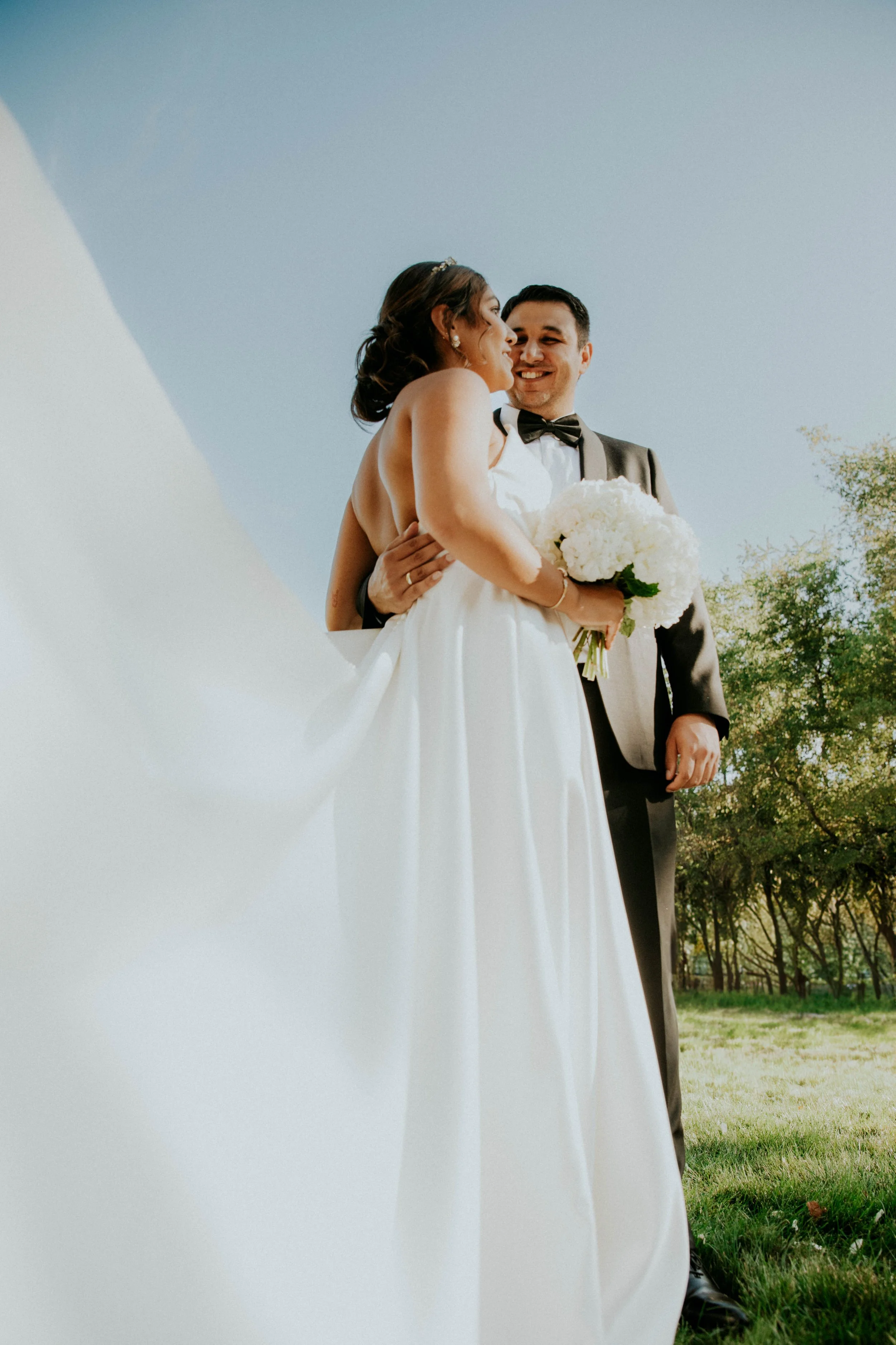 A bride and groom on their wedding day, standing outdoors with a clear sky and trees in the background. The bride holds a bouquet of white flowers, and the groom is dressed in a tuxedo with a bow tie.