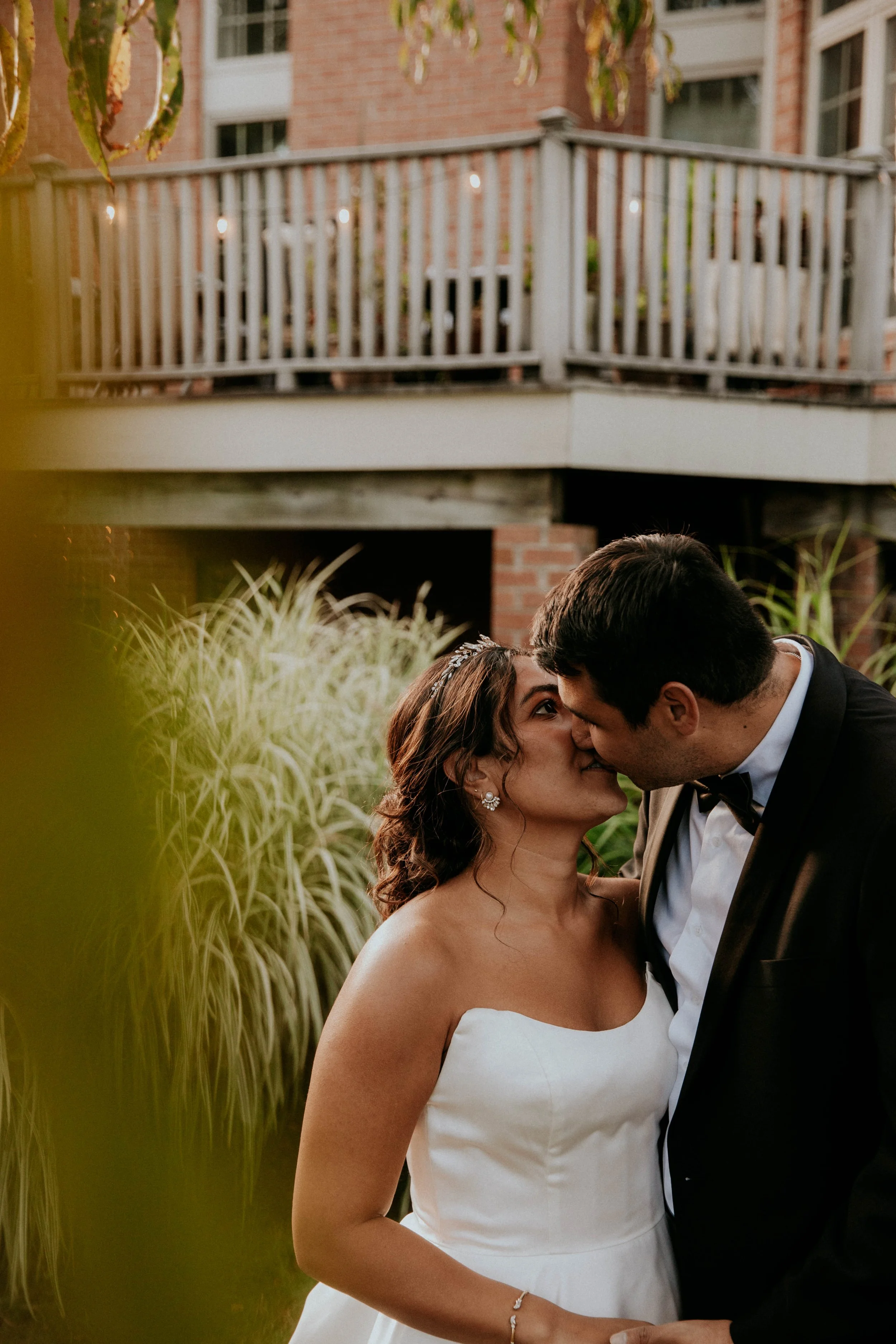 A smiling couple, dressed in wedding attire, sharing a close, intimate moment outdoors with greenery and a brick house with a balcony in the background.