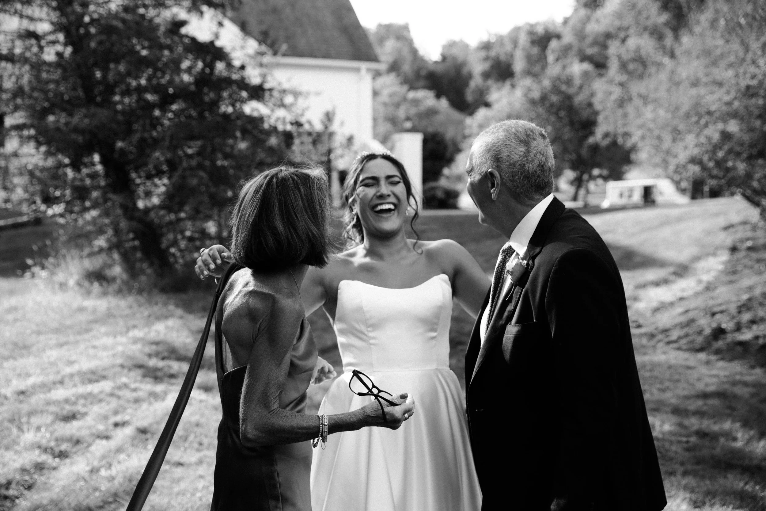 Black and white photo of three people outdoors, smiling and embracing each other, with a house and trees in the background.