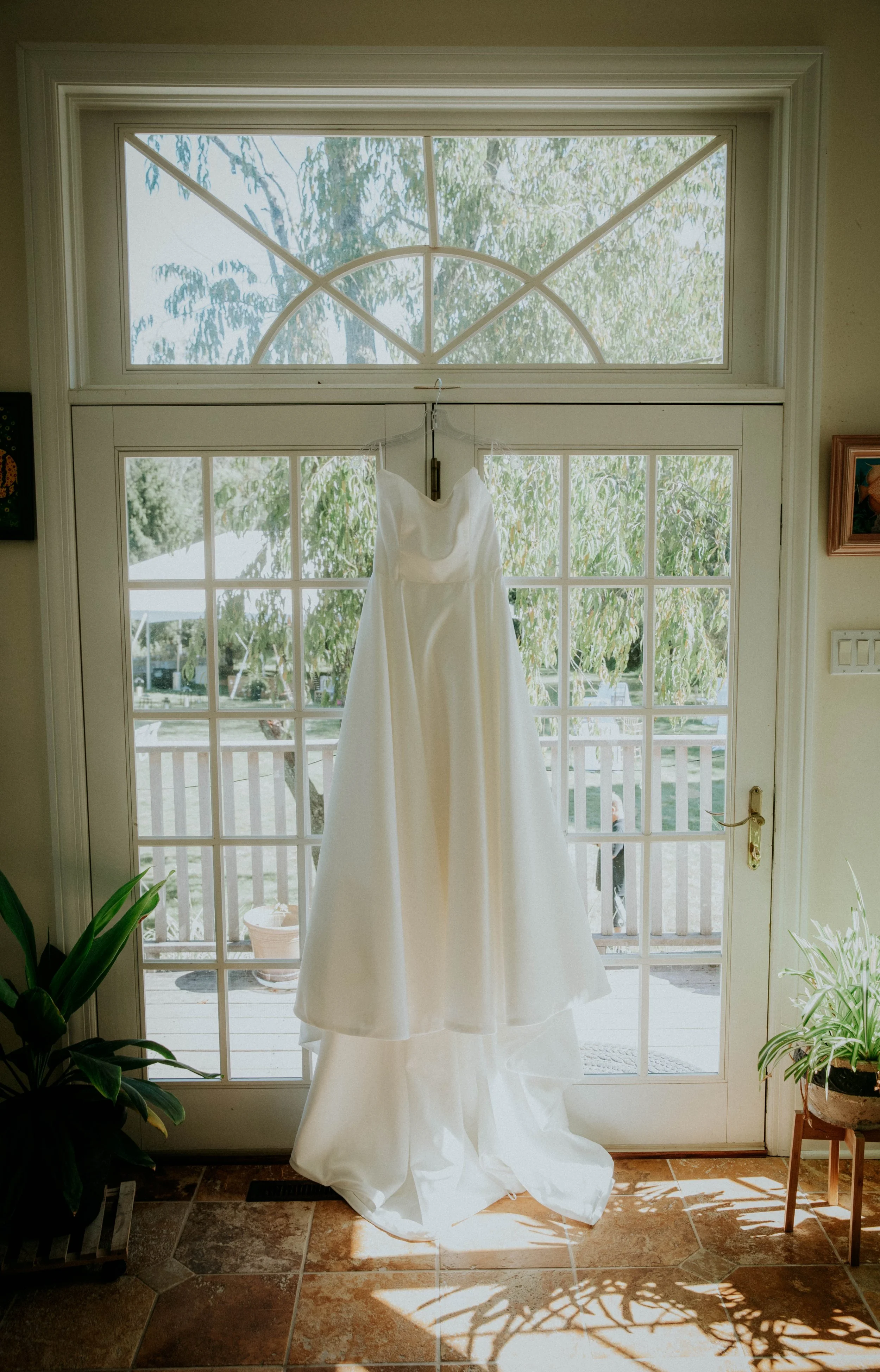 A white wedding dress hanging in front of a large window with a view of trees outside.