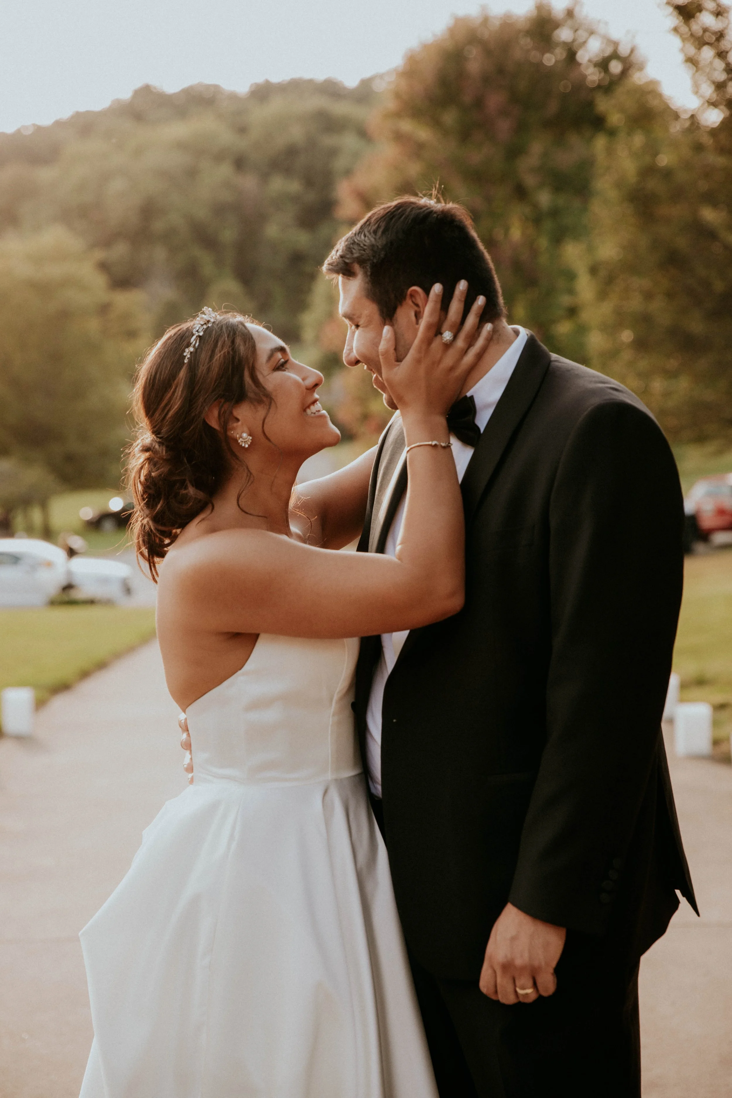A bride and groom sharing a joyful moment outdoors during sunset, with the bride wearing a white wedding dress and tiara, and the groom in a black tuxedo. They are gazing into each other's eyes, smiling, with trees and a few parked cars visible in th