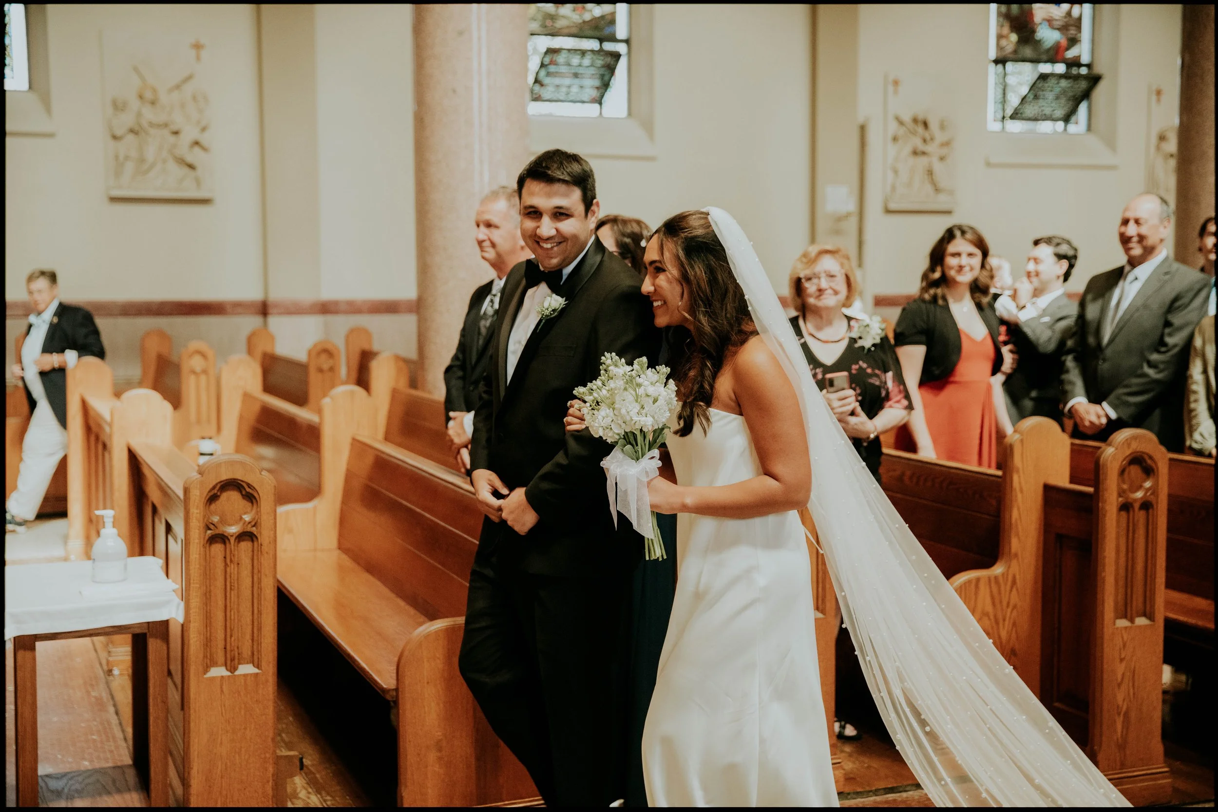 A bride and groom smiling during their wedding ceremony inside a church, surrounded by family and friends.