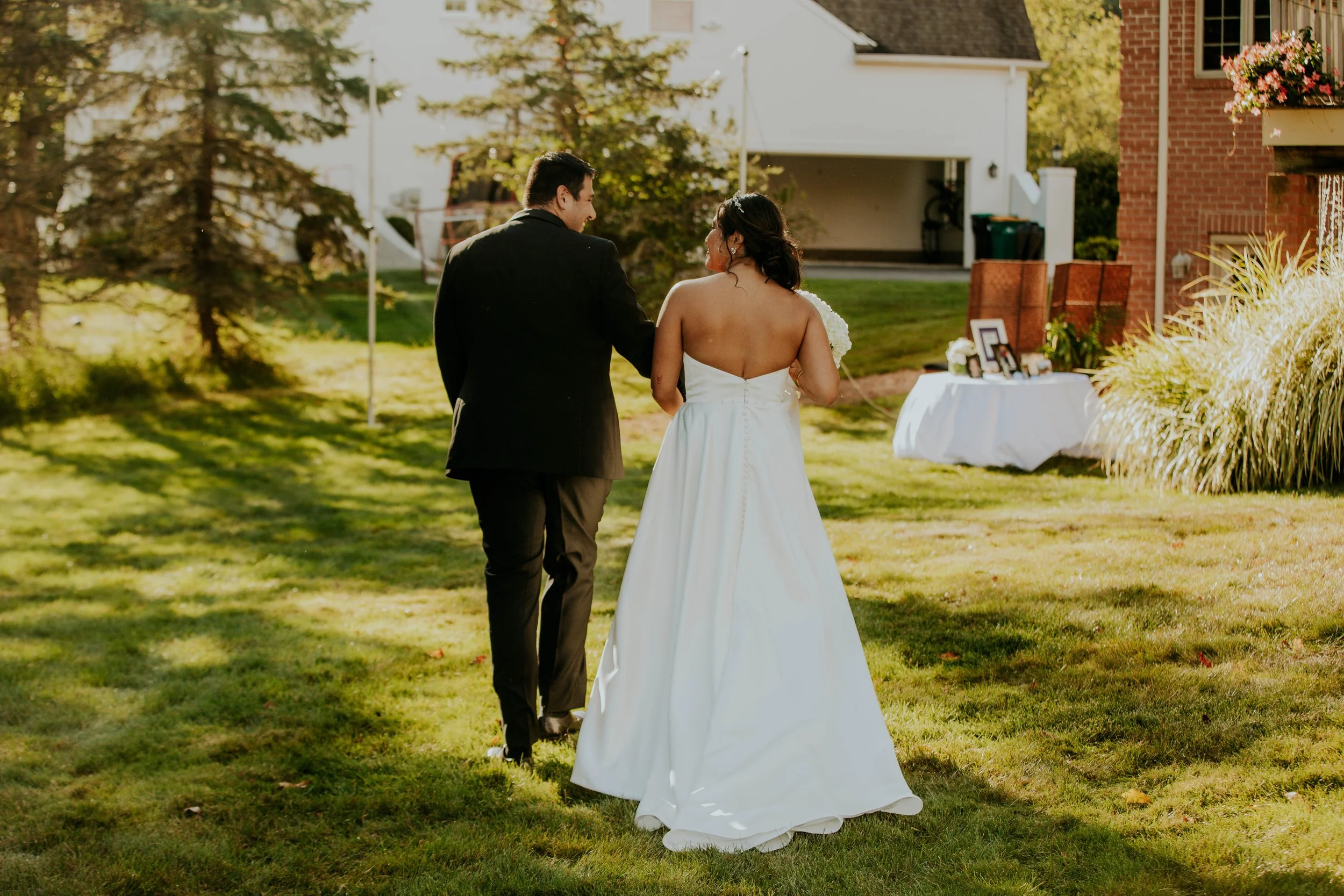 A newlywed couple walking together outdoors on a grassy lawn during late afternoon or early evening, with the bride in a white strapless wedding gown and the groom in a black suit. A table with framed photos and decorations is nearby, and residential