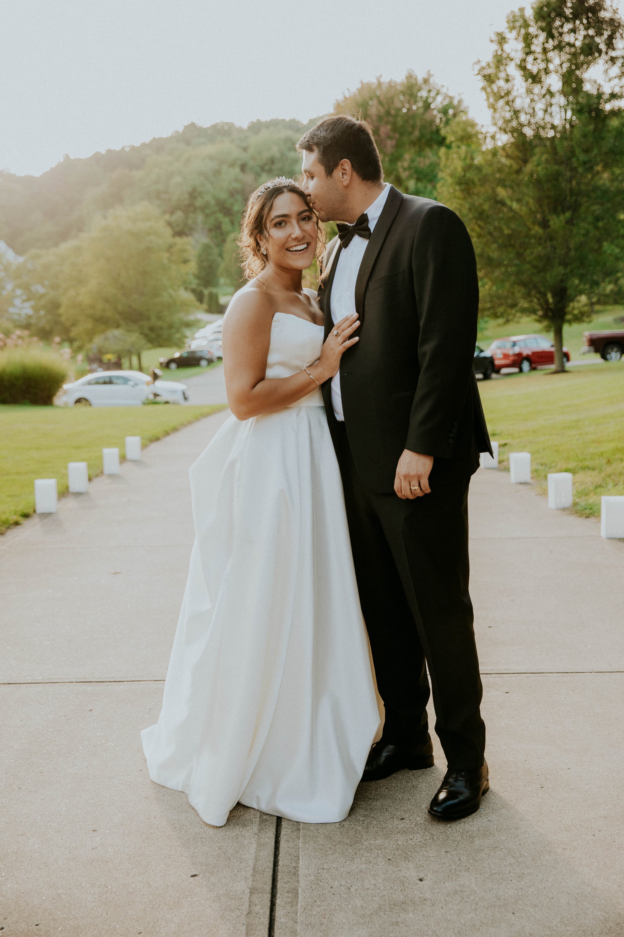 A newlywed couple outdoors at sunset; the bride in a strapless white wedding gown with a tiara, smiling at camera; the groom in a black tuxedo kissing her forehead.