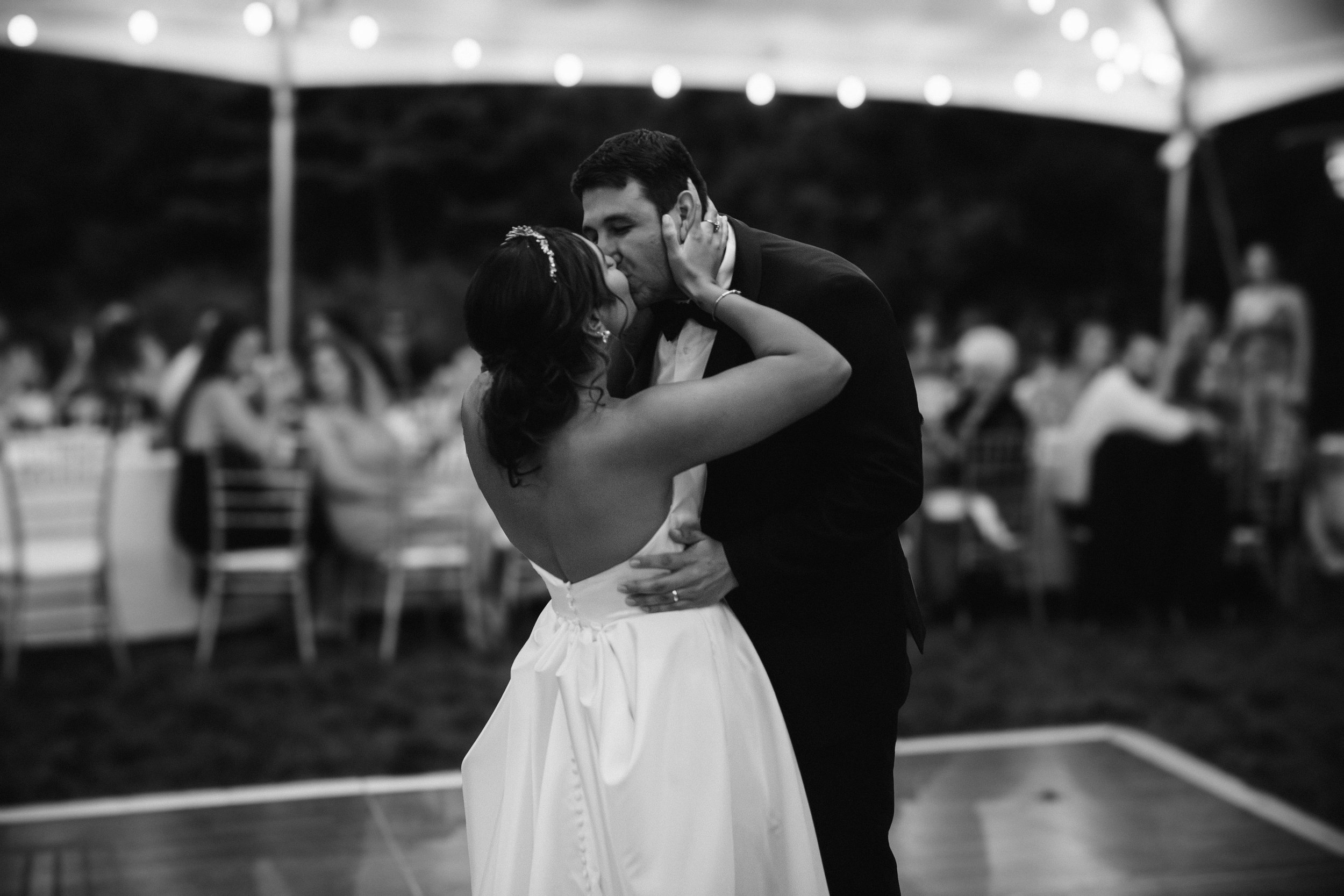 A couple dancing closely at a wedding reception, with the bride in a strapless gown and the groom in a tuxedo, surrounded by blurred guests and hanging string lights.
