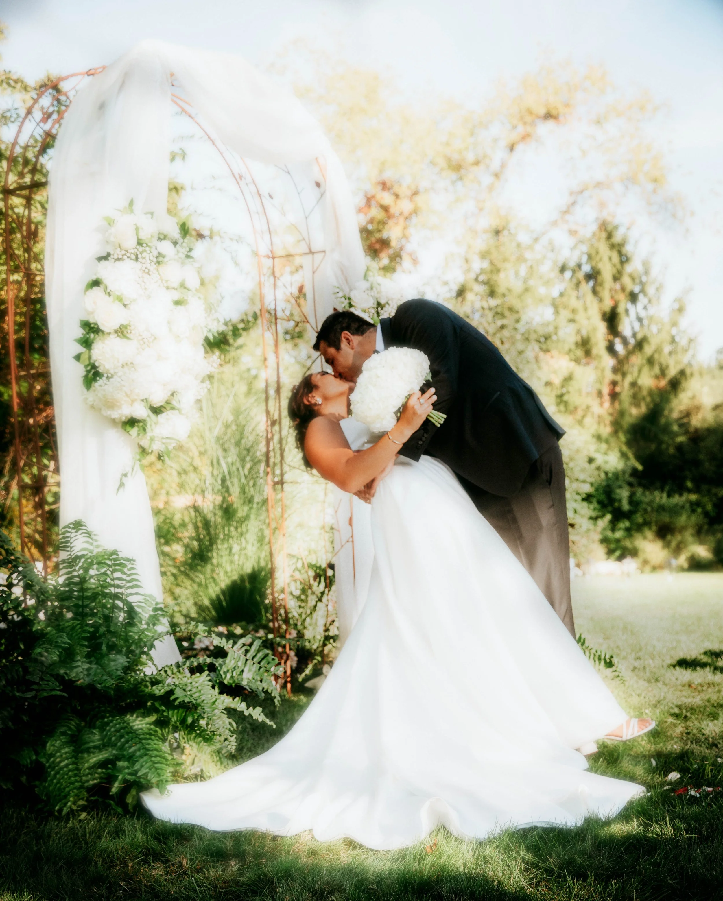 A bride and groom share a kiss outdoors during their wedding ceremony, with the bride wearing a white gown and holding a bouquet of white flowers, and the groom in a black suit. They are under a decorated arch with white flowers and greenery, set in 