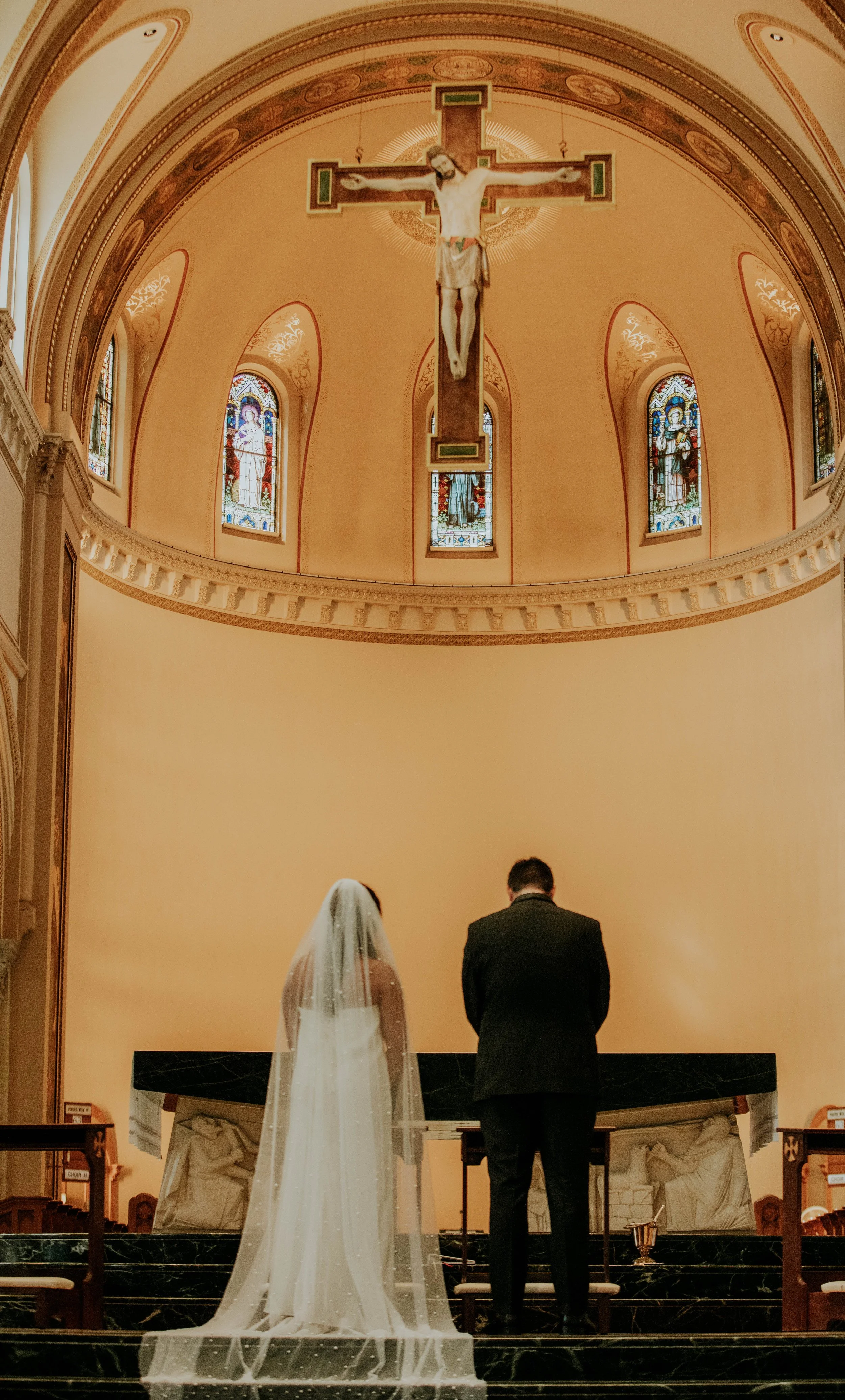 A bride and groom stand together at the altar inside a church, with their backs facing the camera. The bride is wearing a long veil and gown, while the groom is dressed in a dark suit. Above them is a large crucifix of Jesus Christ hanging from the c