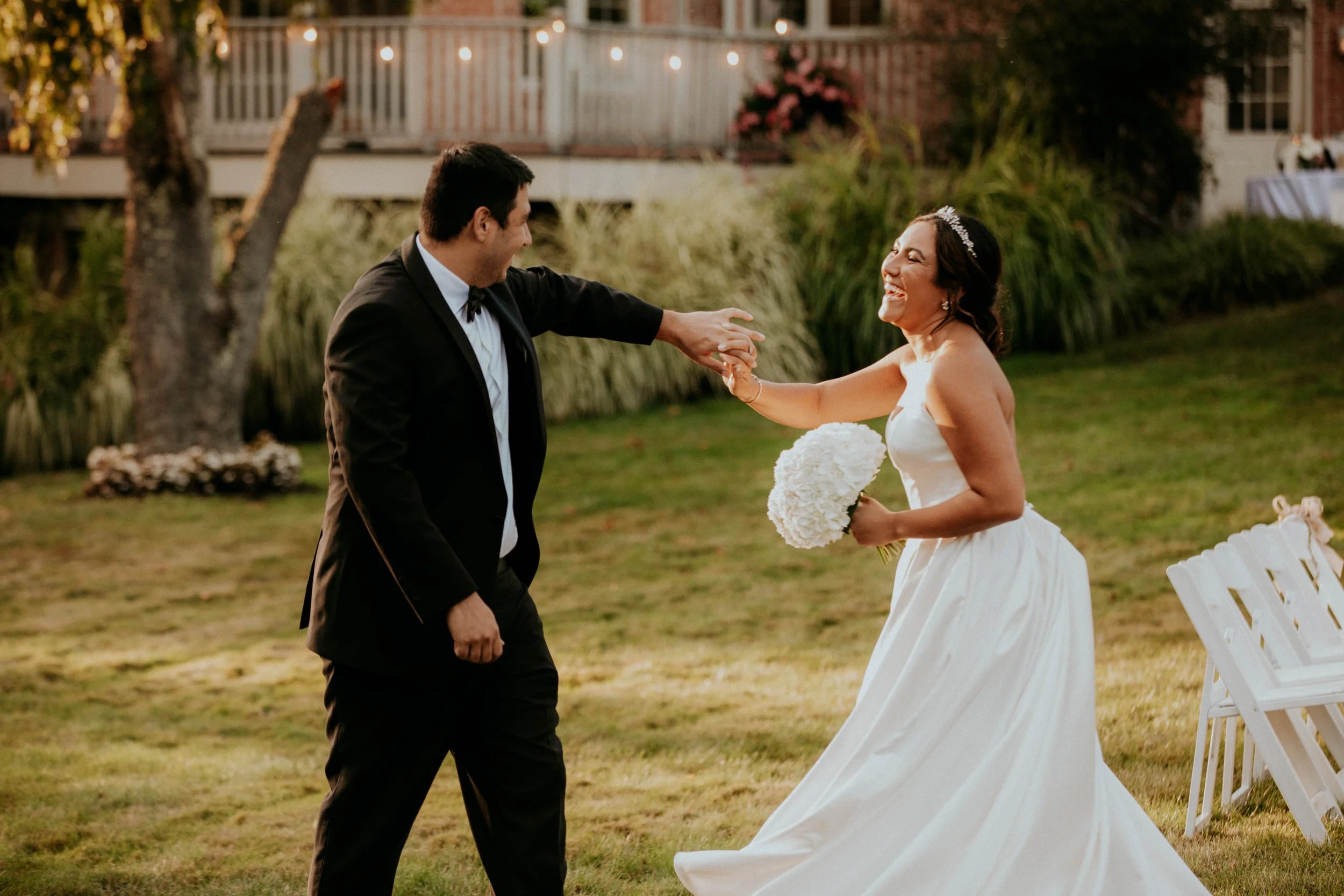 A newlywed couple smiling and holding hands at an outdoor wedding reception during sunset, with string lights and a decorated building in the background.