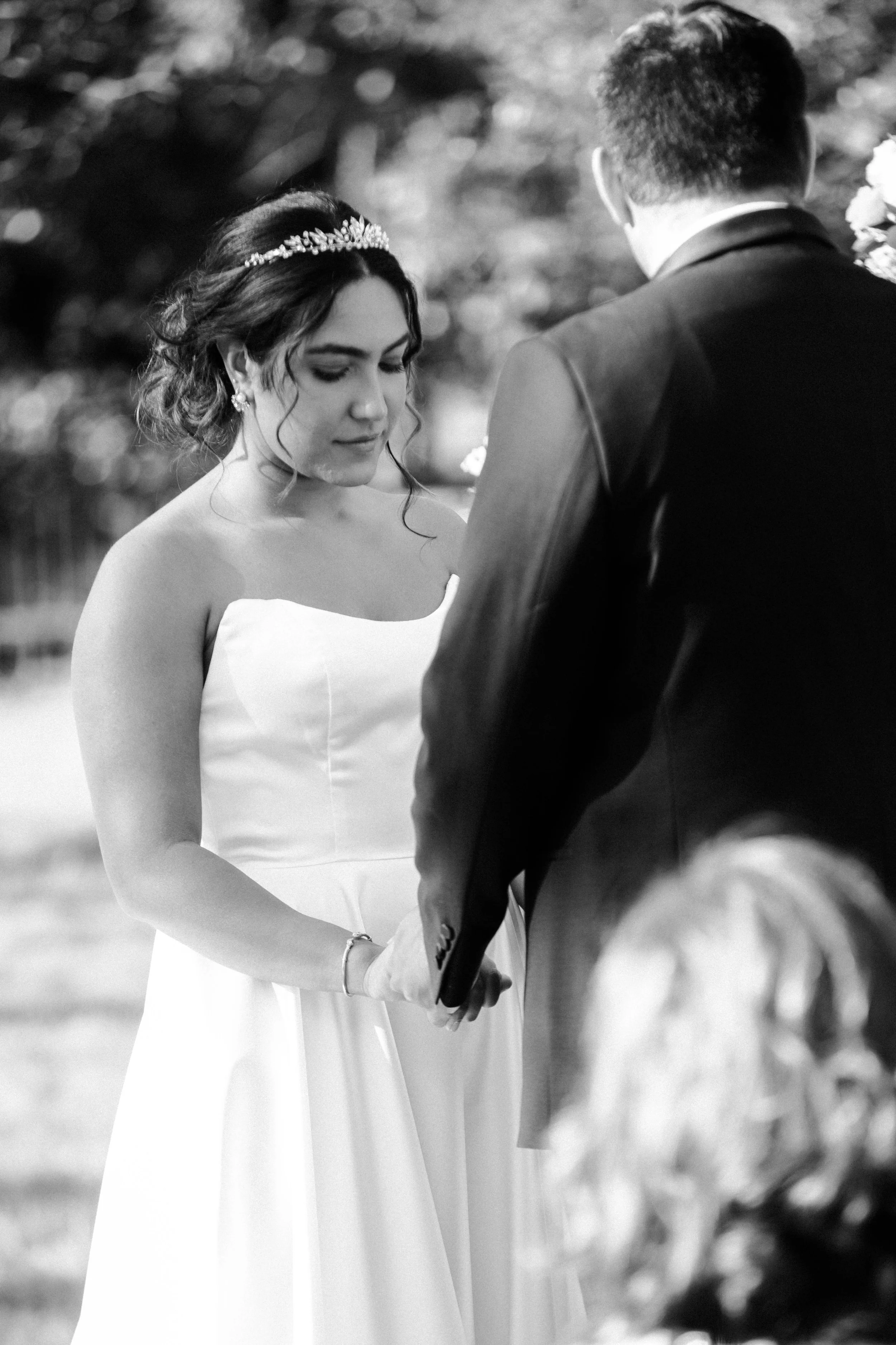 A black-and-white photo of a bride and groom holding hands during their wedding ceremony outdoors. The bride is wearing a strapless wedding dress with a tiara and earrings, and has dark, wavy hair. The groom, seen from the back, is wearing a dark sui