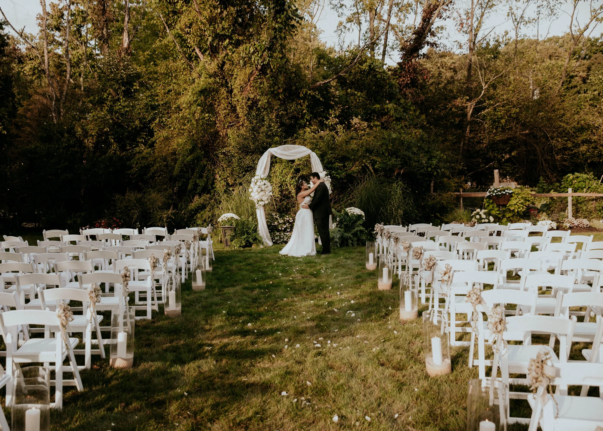 A couple standing under a wedding arch decorated with white flowers, surrounded by white chairs with flower decorations, set up for an outdoor wedding ceremony in a wooded area.