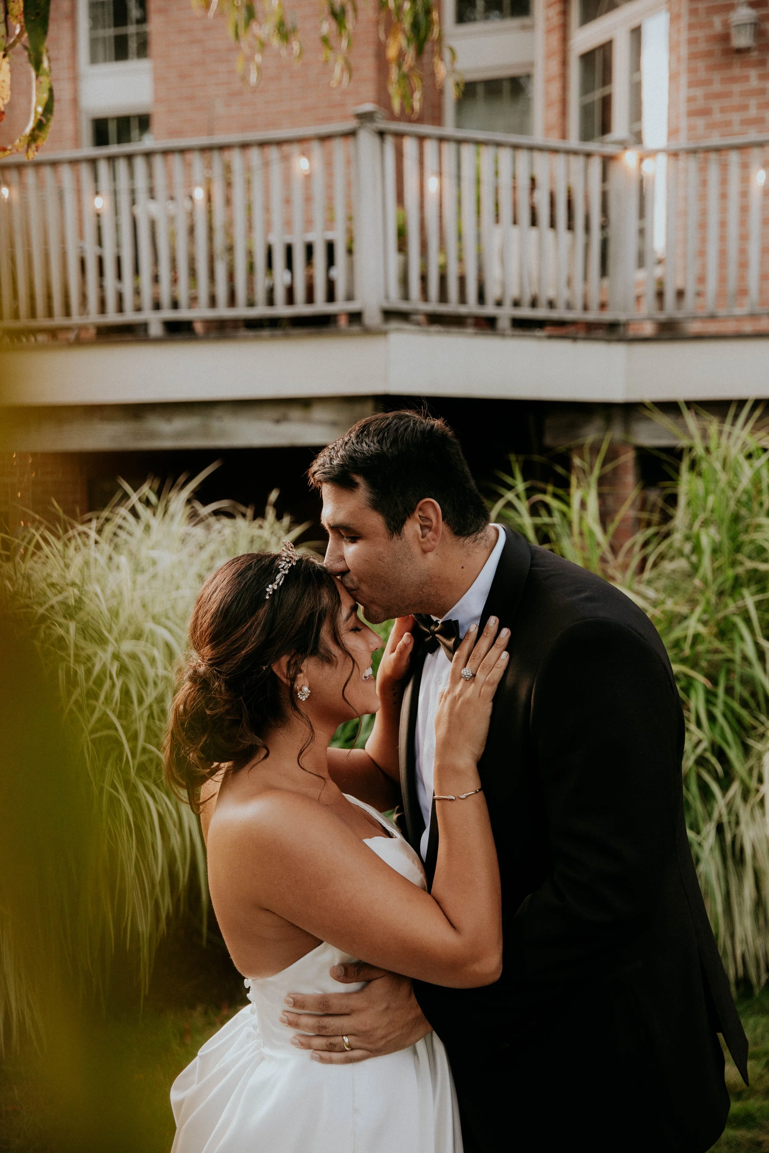 A bride and groom embracing each other outdoors, with their eyes closed, sharing a tender moment on their wedding day, surrounded by greenery and a brick building in the background.