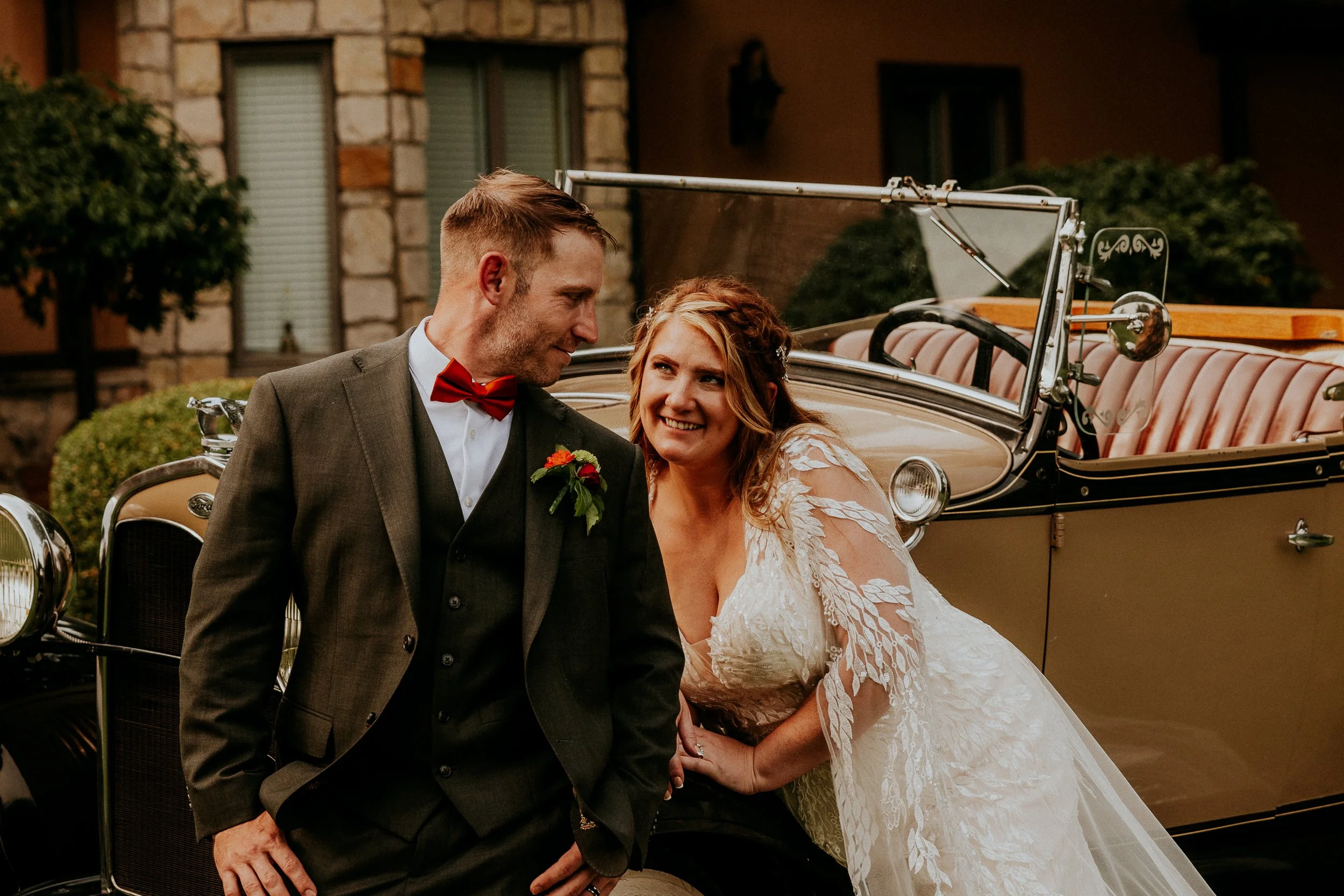 A bride and groom smiling and looking at each other, standing in front of a vintage car during their wedding celebration.