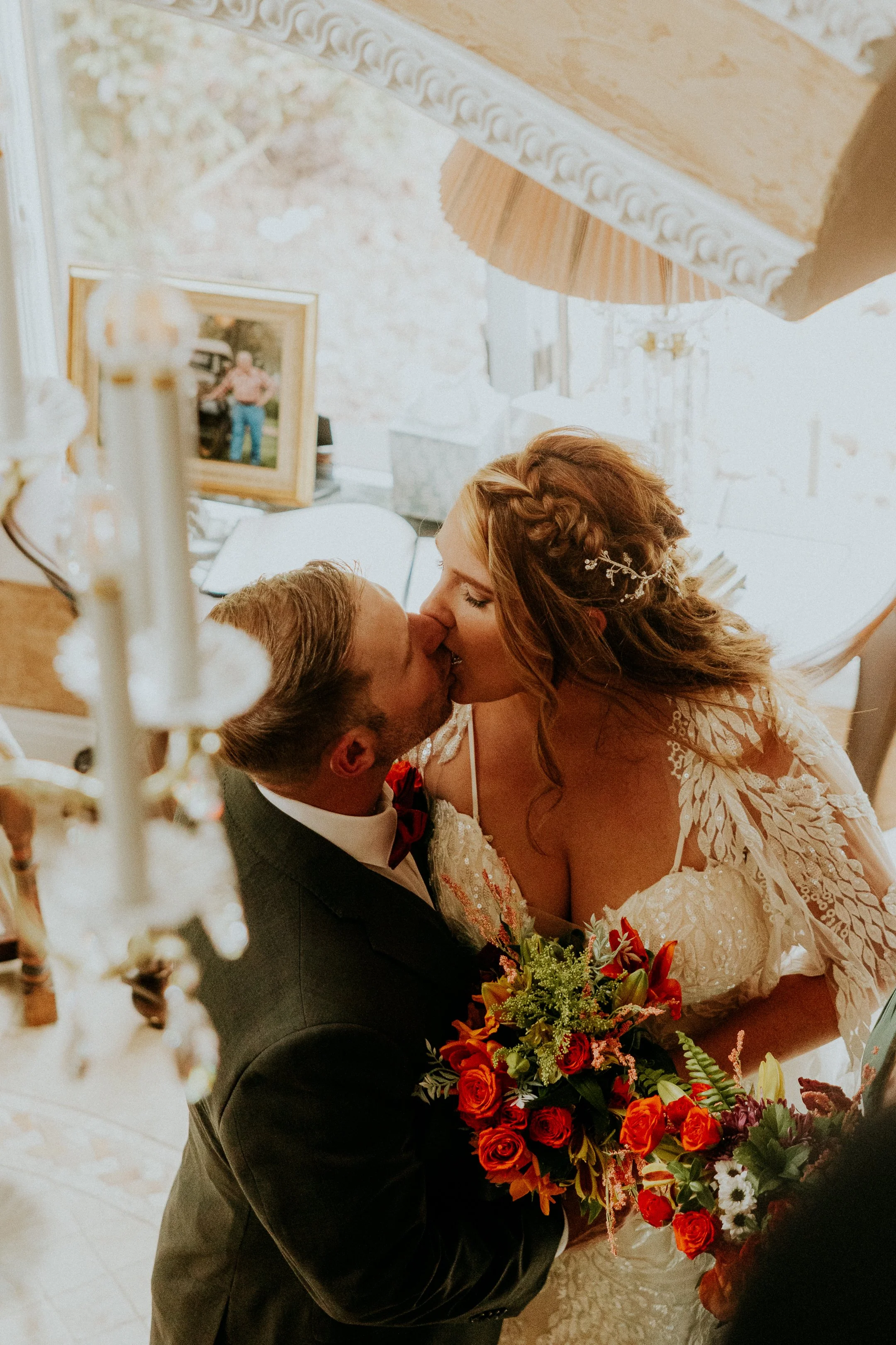 A bride and groom share a kiss at their wedding, with the bride holding a colorful bouquet of flowers, inside a decorated room.