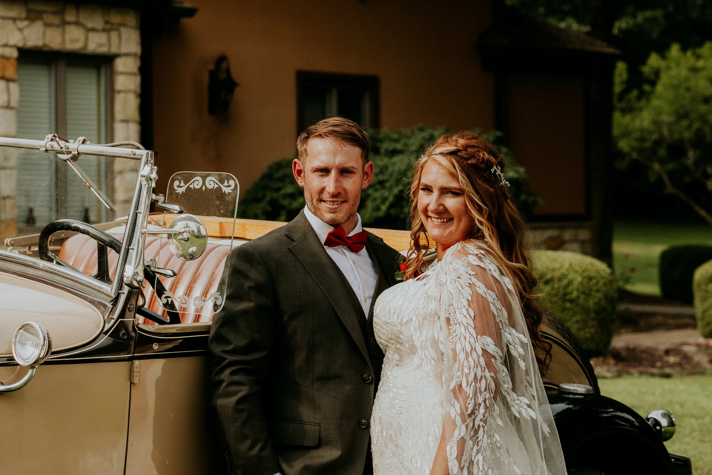 A newlywed couple dressed in wedding attire standing beside a vintage car outdoors. The groom is in a dark suit with a red bow tie, and the bride is in a white lace wedding dress with her hair styled in loose waves with a small braid. The background 