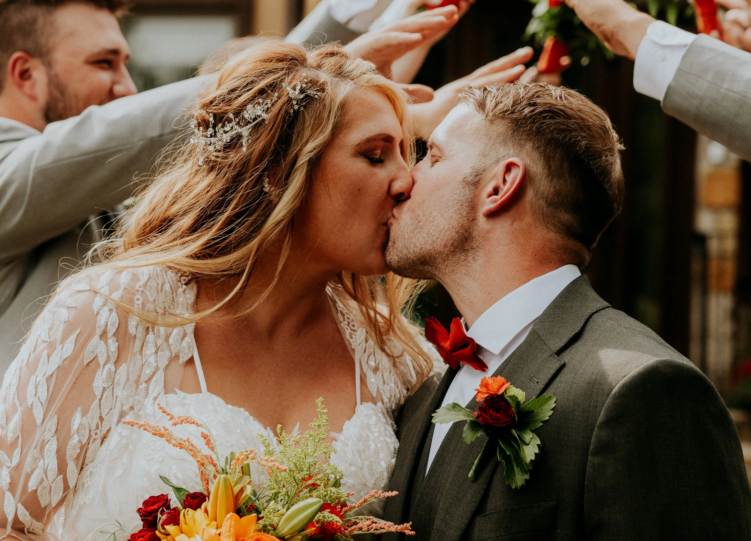 A couple sharing a kiss at their wedding ceremony, with the bride holding a bouquet of flowers, and the groom wearing a suit with a boutonniere. Two other people in the background reach out towards the couple.