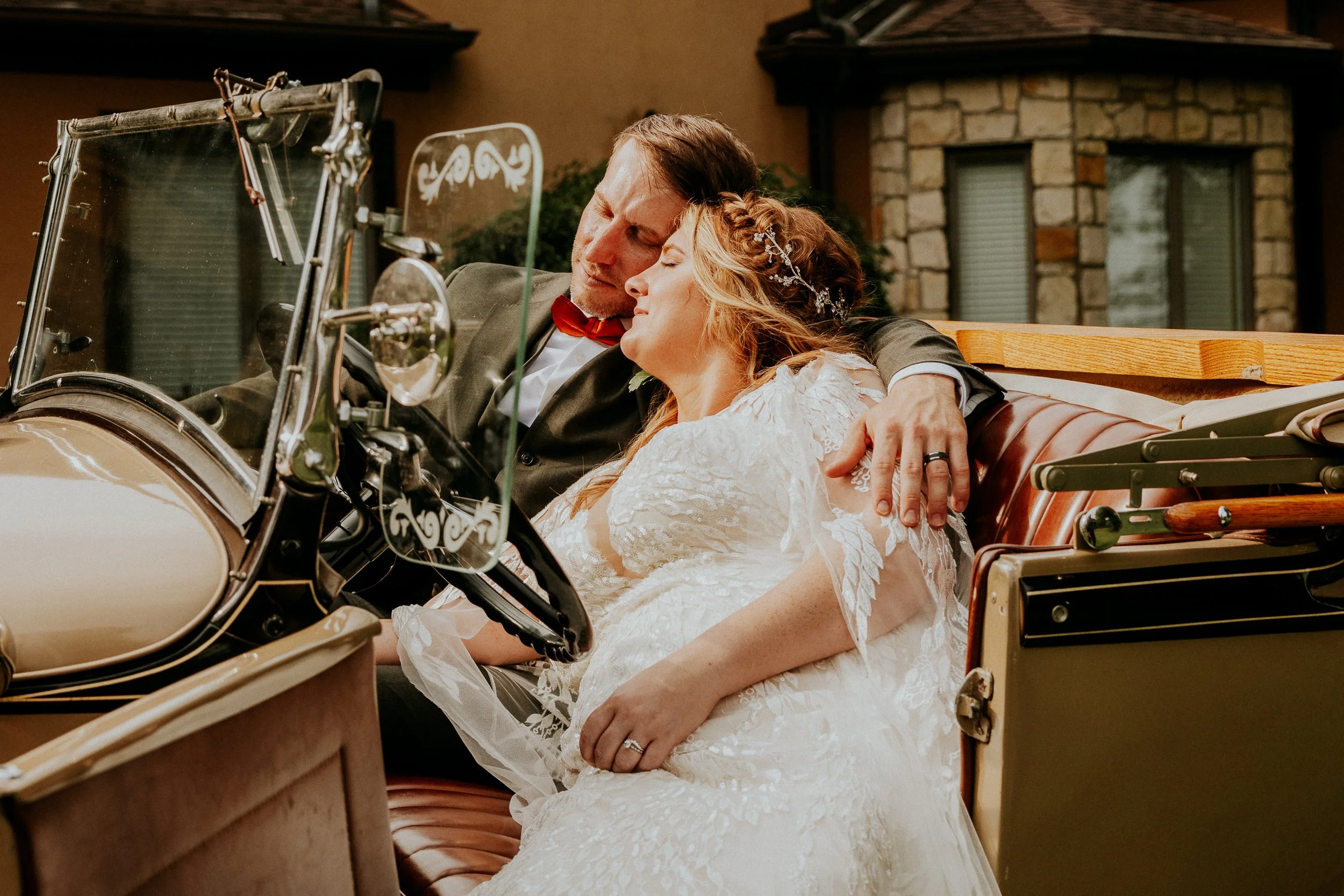 A bride and groom sitting in a vintage car, embracing and resting their heads together.